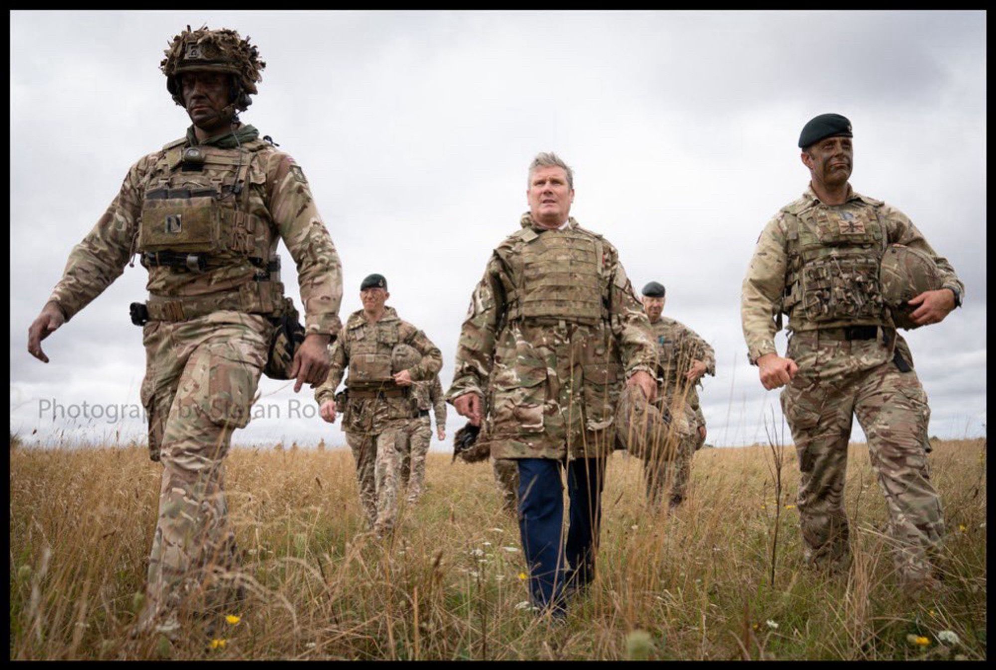 Keir Starmer wearing military battle dress over his suit, looking tiny amongst a group of soldiers