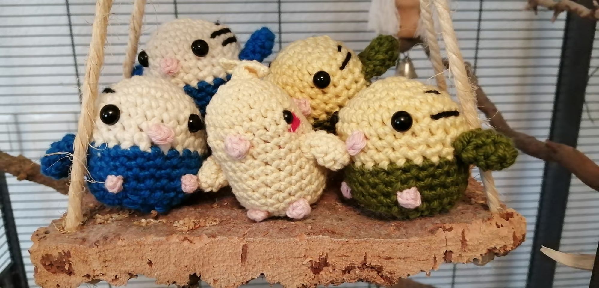 A photo of little crocheted borbs-budgies, and two kinds of cockatiels-posed on a hanging bird perch/swing inside a bird cage. Photo belongs to, and borbs created by, Aucraptor of Bluesky