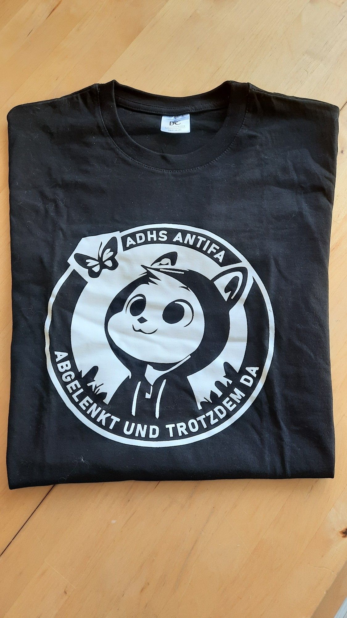 A black t-shirt with a comic illustration of a cat wearing an hoodie and being distracted by a butterfly. The text on the t-shirt days ADHS Antifa distracted and but still there.