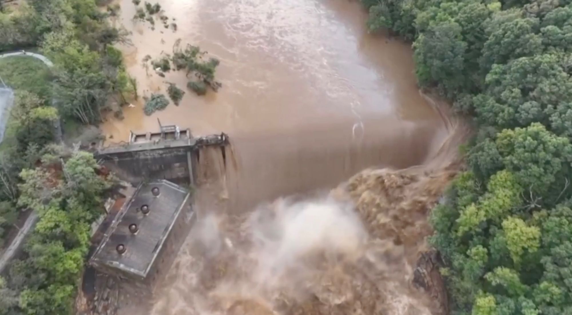 Photo from TVA of Nolichucky Dam, with brown river raging over the top of it and around it. Part of the river looks to be going around the dam, with waves higher than a building next to the dam.