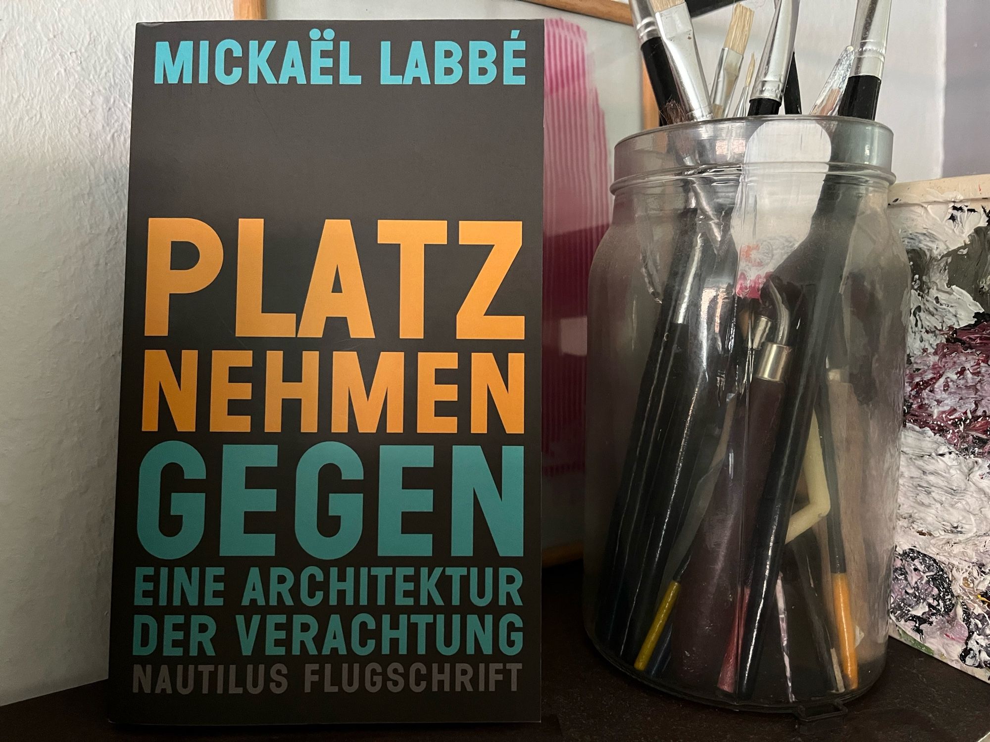 Buch „Platz nehmen - gegen eine Architektur der Verachtung“ im Regal neben einem Glas voller Pinsel.