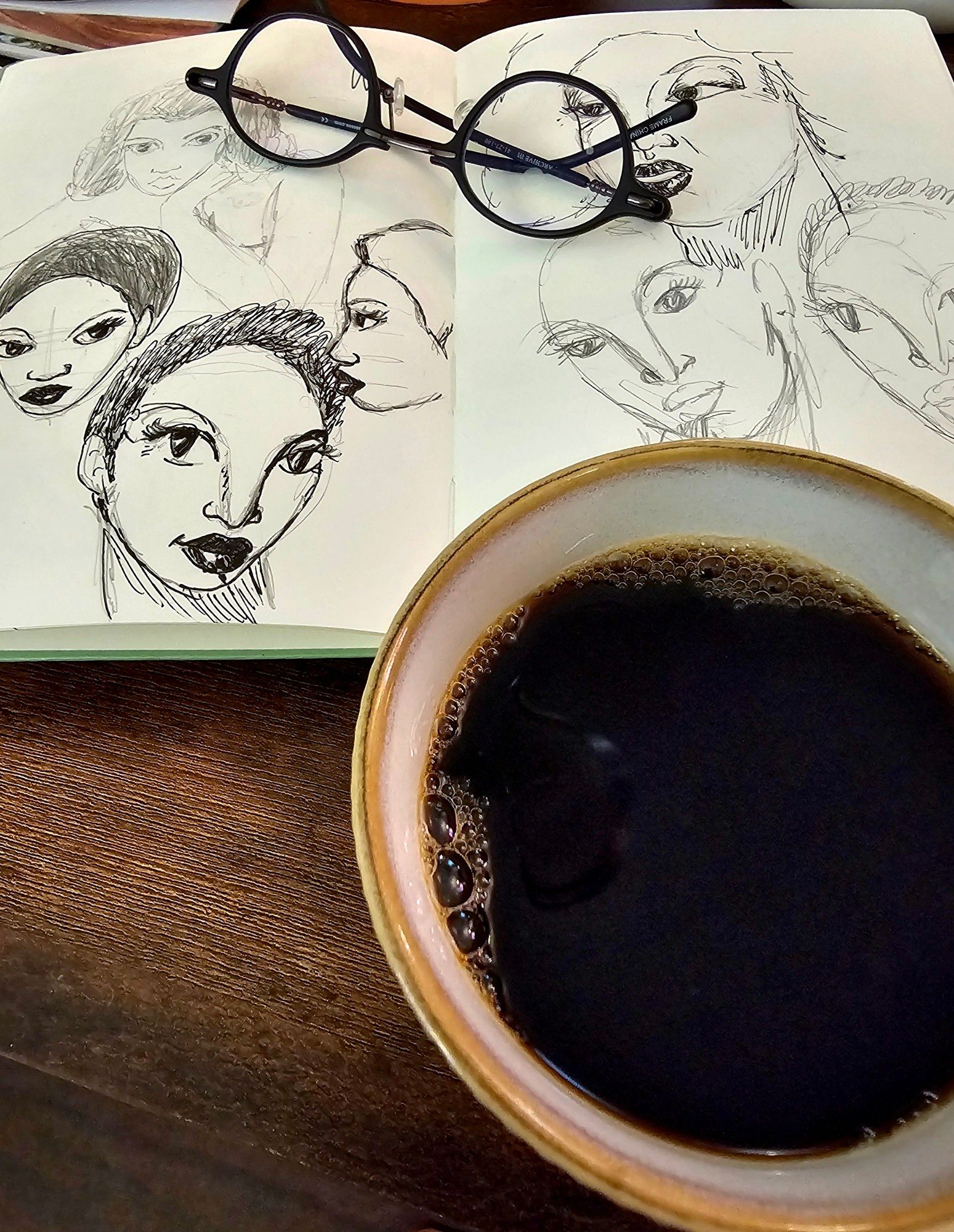 photo of black coffee and an open notebook full of sketched feminine heads with short hair.