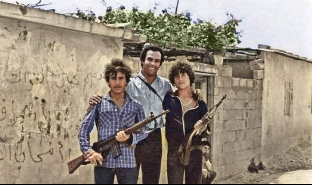 Huey P. Newton has his arms around two young Palestinians holding rifles while smiling. In the background is a building of some sort in rough shape, with a tree partially obscured by it. The brickwork is missing a section in one spot. It is a building in an unknown refugee camp. There appears to be some kind of writing on the one wall, but it is largely washed off and damaged, rendering it likely illegible.