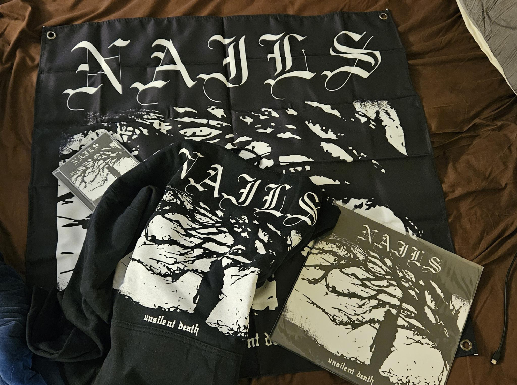 Nails - Unsilent Death flag, on top of which is a Nails - Unsilent Death kangaroo jacket, a Nails - Unsilent Death vinyl (first pressing on Streetcleaner), Nails - Unsilent death CD (2013 re-release on Southern Lord).

Not pictured: FLAC version of the 2020 10th Anniversary Edition with bonus tracks