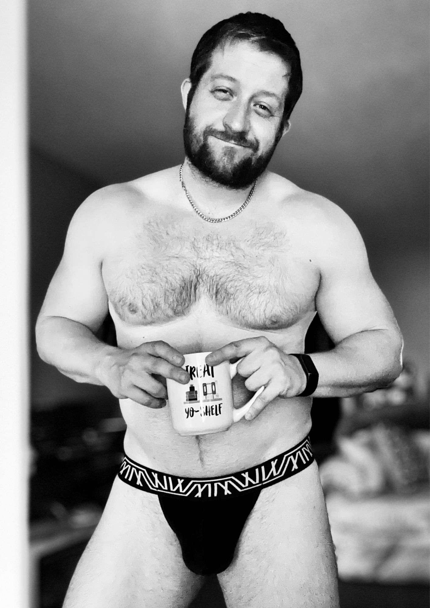 Black and white photo of a gay man standing with a smirk that disguises his physical aches and pains. He’s drinking coffee from a mug featuring a “Treat Yo-Shelf” pun, beloved by both book nerds and “Parks and Rec” fans alike. He’s also wearing one of his partner’s thongs, but the back view remains a coy mystery.