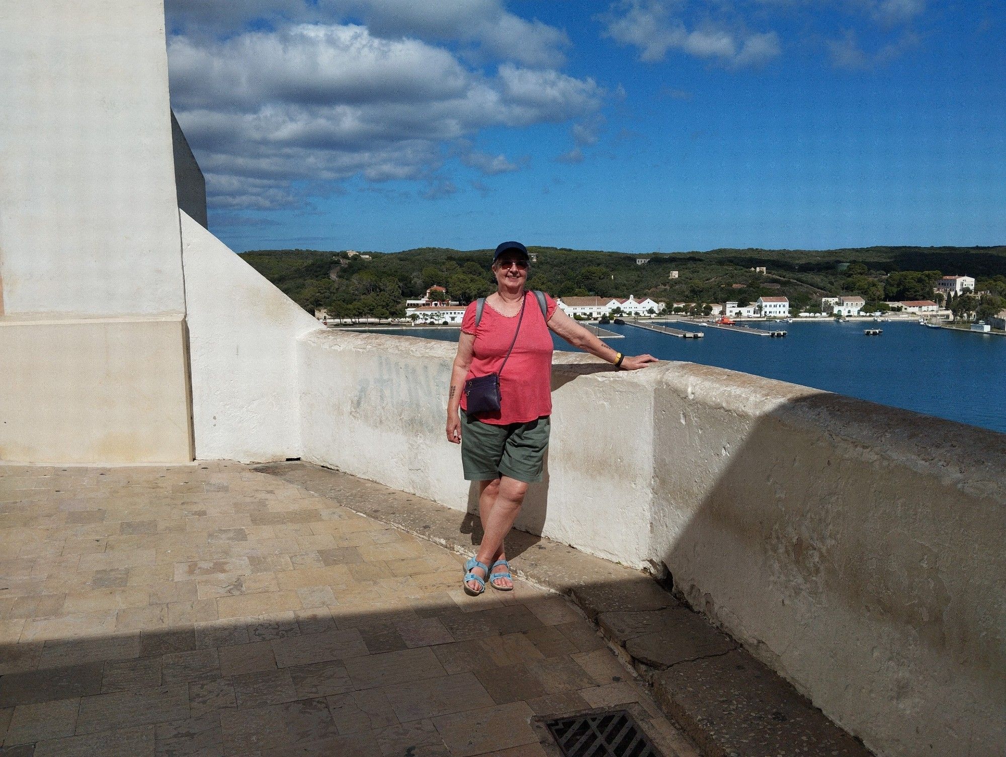 A person in a navy Lacoste cap standing by a white wall overlooking the sea in Mahon