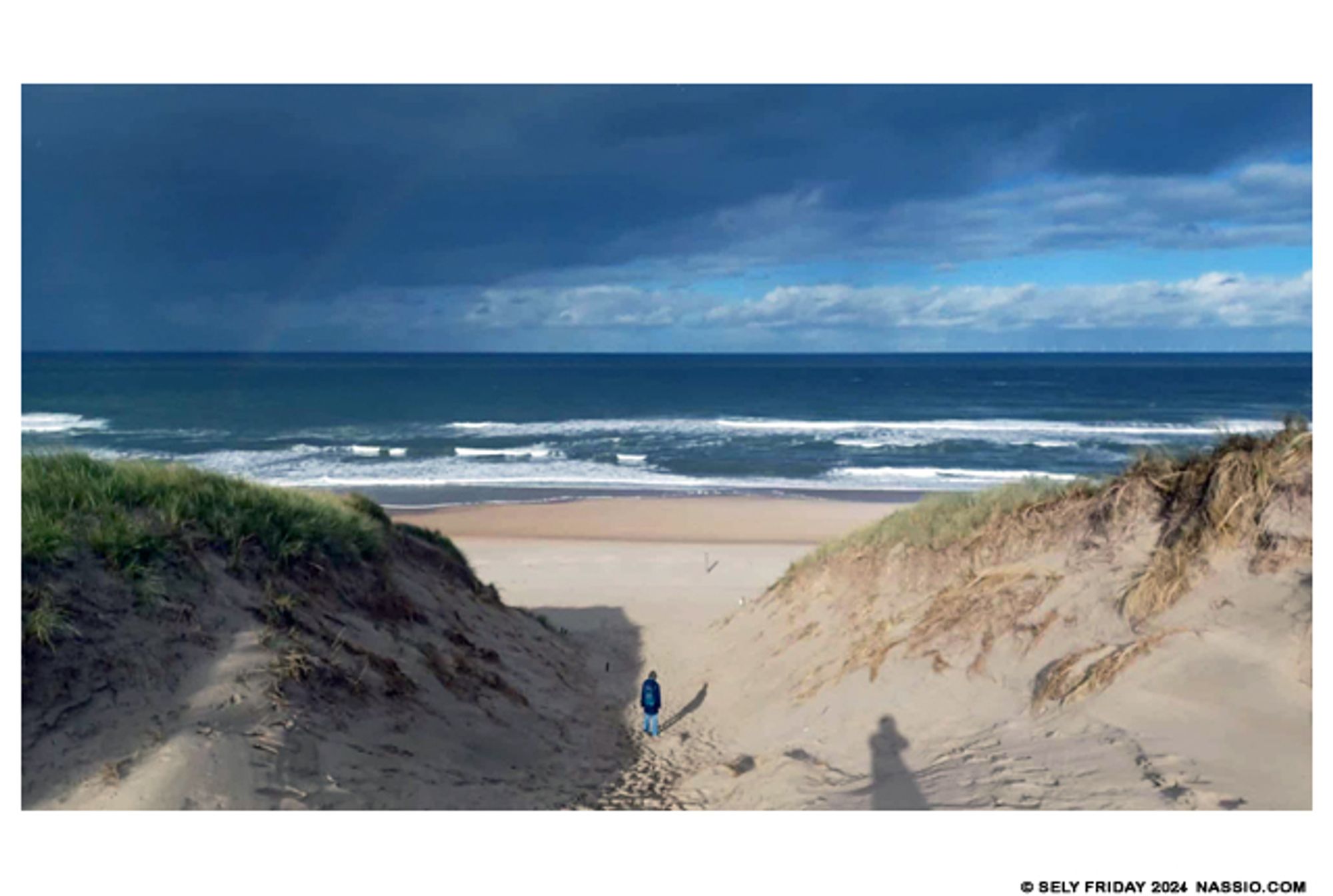 Colour digital photograph of a lady walking down a step channel between two large dunes toward a beach.