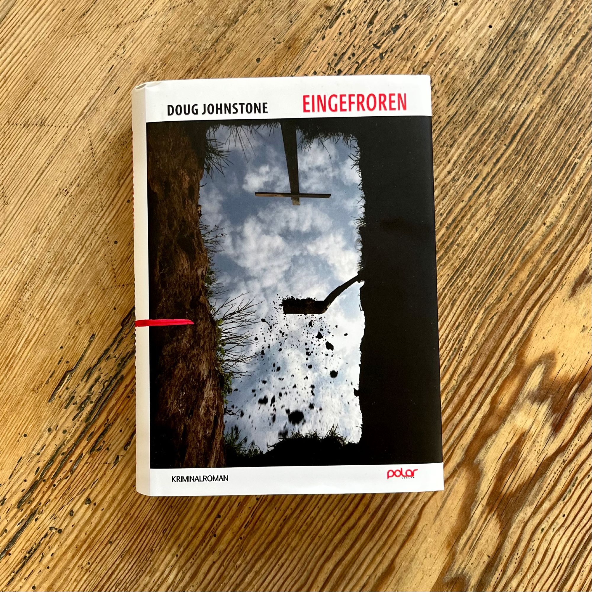 Hardcover auf Holztisch: Doug Johnstone: Eingefrorenm Polar Verlag. Das Cover zeigt die Perspektive von jemanden, der in einem offenen Grab zugeschaufelt wird, Erde rieselt von der Schaufel