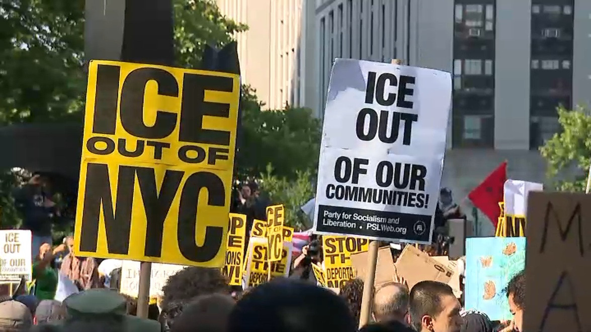 The image captures a protest with numerous signs and people advocating for the removal of ICE (Immigration and Customs Enforcement) from specific locations. The foreground features two prominent signs. The first, on the left, is a bright yellow sign with bold black lettering that reads "ICE OUT OF NYC." To the right of it, a white sign with black lettering states "ICE OUT OF OUR COMMUNITIES!" and includes the designation "Party for Socialism and Liberation. PSLWeb.org."

Behind these signs, a diverse crowd of people can be seen holding other signs, some of which are partially visible. One sign mentions "STOP THE DEPORTATIONS!" and another displays a picture of butterflies. A red flag is also visible, adding to the atmosphere of activism and political engagement.

In the background, a large building with multiple windows is visible, providing a sense of location and scale to the protest. The overall composition of the image conveys a sense of urgency and purpose, as individuals come together to express their opposition to ICE and advocate for change within their communities.