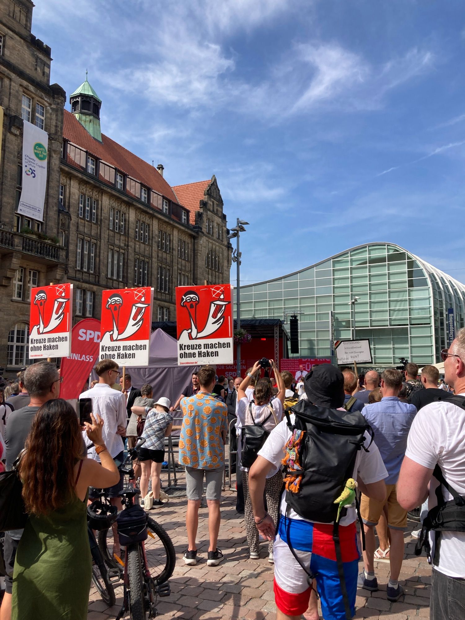 Chemnitzer Neumarkt, im Hintergrund das Peek und Cloppenburg-Gebäude, links das Rathaus. Der Bundeskanzler spricht vorne auf der Bühne, eine Menschenmenge steht davor. 3 Personen halten Schilder von Storch Hainar: “Kreuze machen ohne Haken”
Im Hintergrund dürfen die Freien Sachsen so laut skandieren, dass der Bundeskanzler kaum zu hören ist.