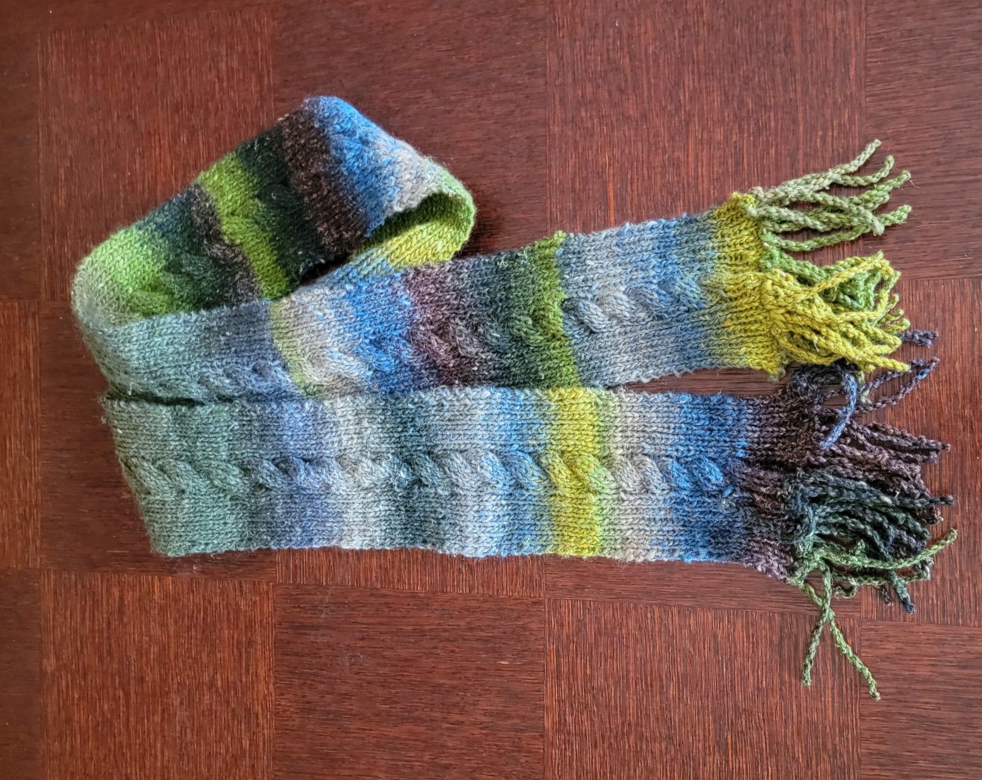 Scarf with a single center cable and fringes in gray, blue, green, purple variegated noro yarn on a wood background.