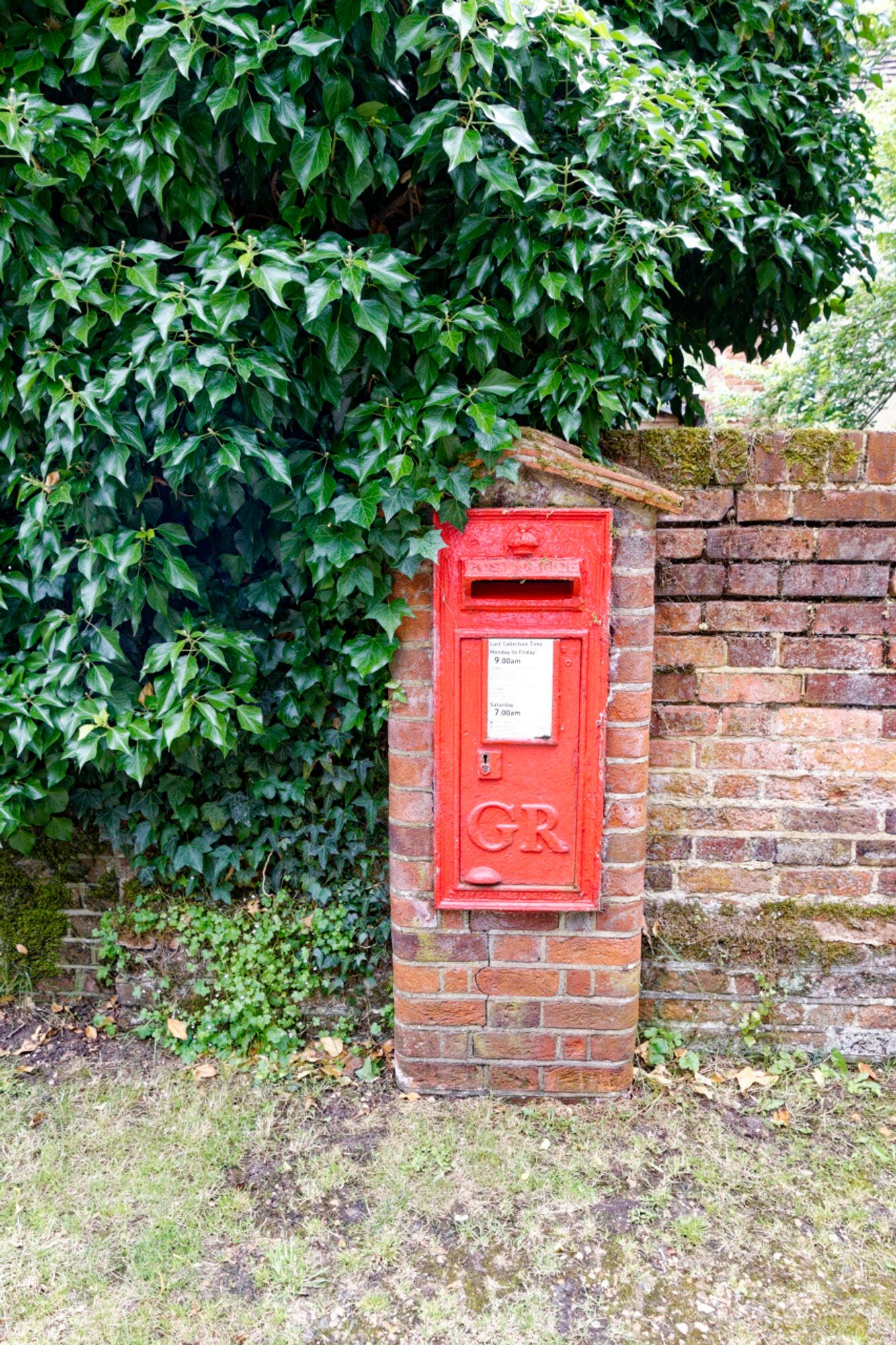 A red postbox with cypher GR sits in a brick wall with a little covering resembling a hat. There is green foliage on most of the left side of the photo.