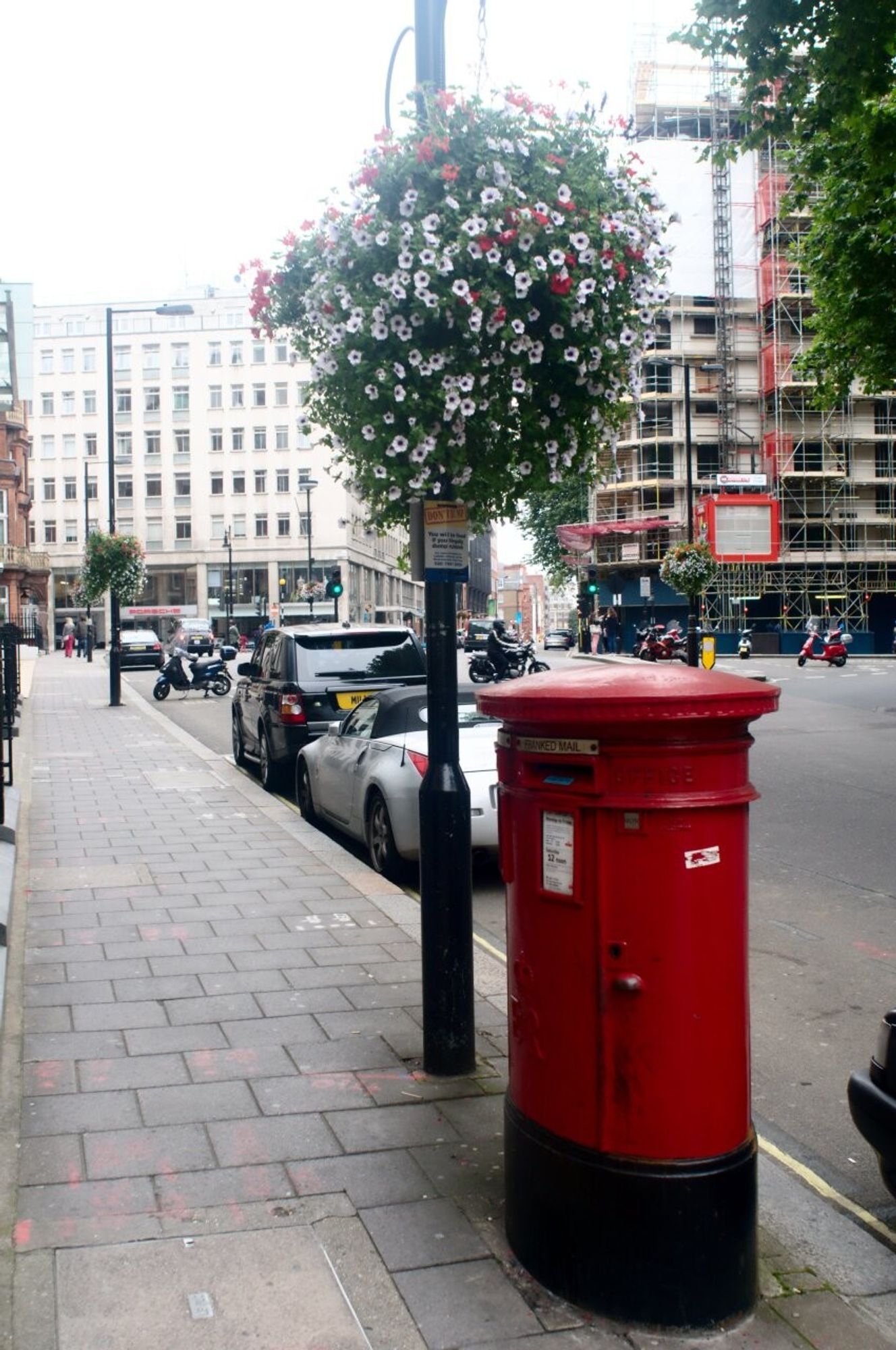 An oval-shaped pillar postbox sitting on the pavement. Next to it is a lamp post with a colourful ball of flowers.