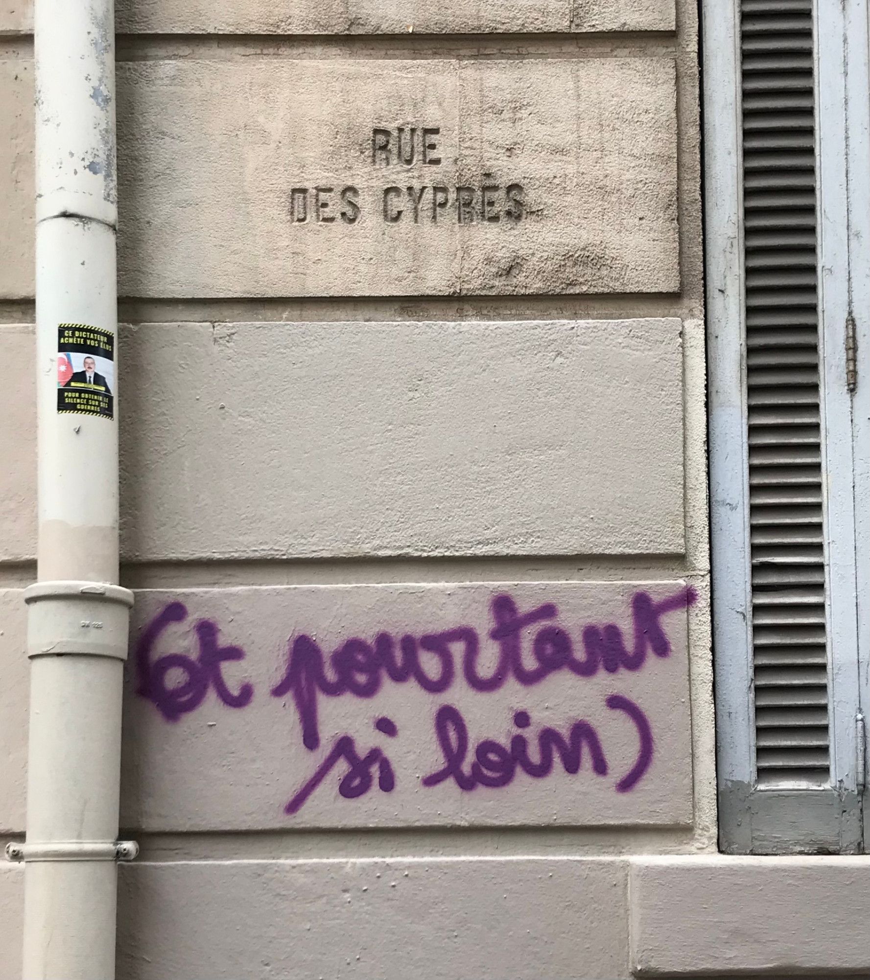 Sur un mur est gravé le nom de la rue "rue des cyprès" et dessous un tag violet qui dit "et pourtant si loin"