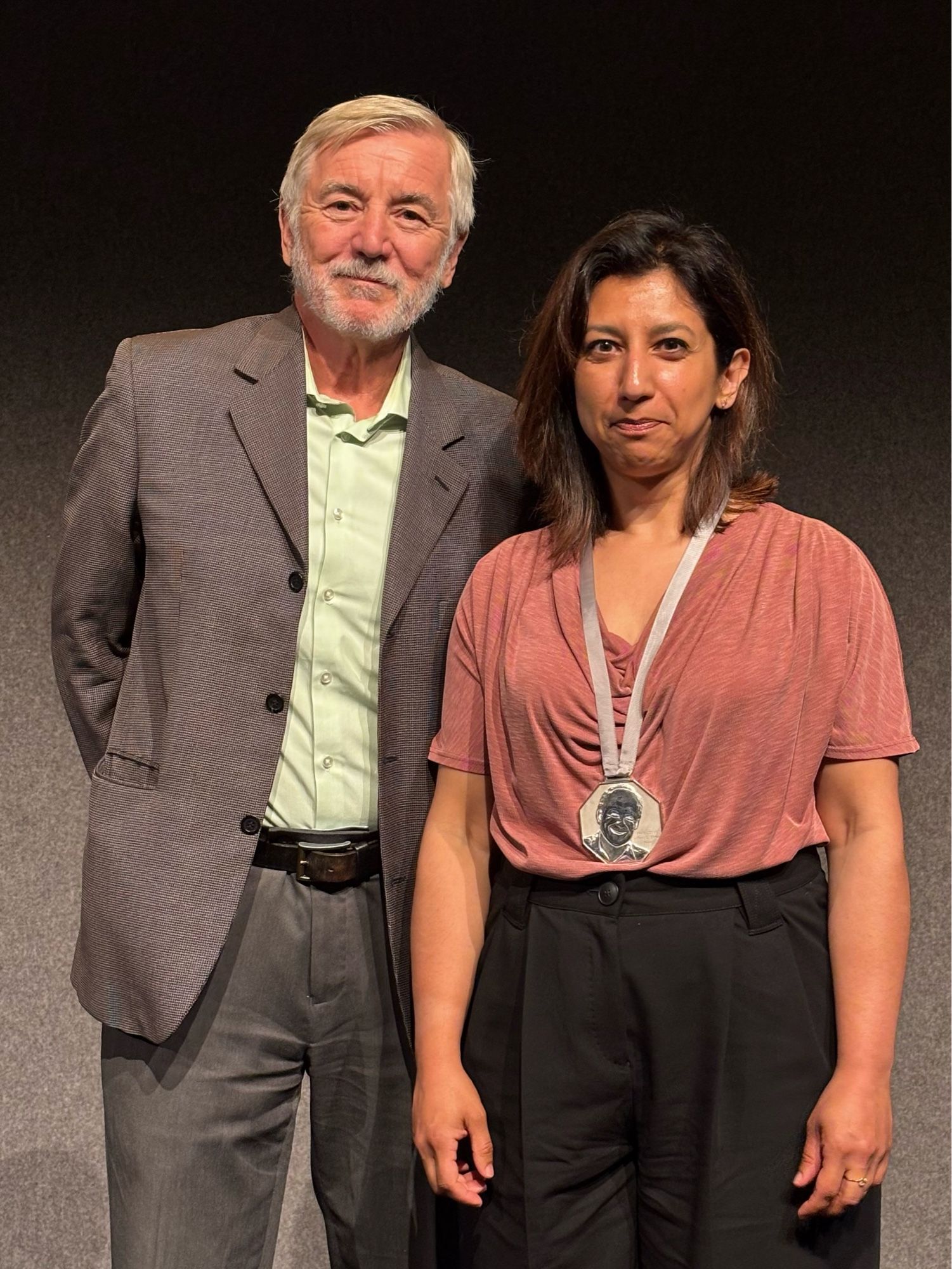 Asifa Majid and Bob Glushko are standing together against a dark background. Asifa, on the right, is wearing a pink shirt and black pants, with a silver medal around her neck (the Elman Prize Medal).