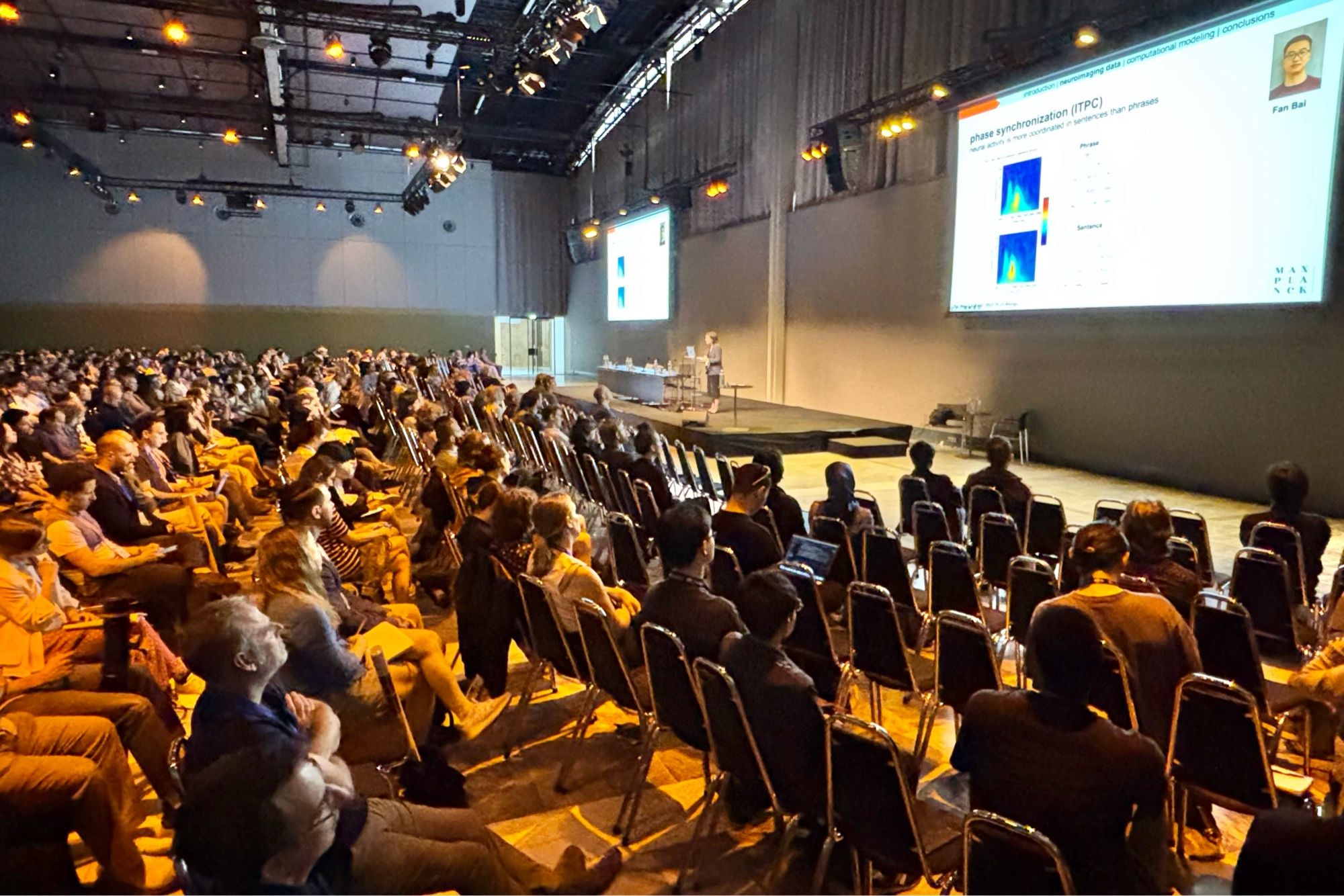 A large audience is seated in a conference room, attentively watching a presentation. The presenter - Andrea E. Martin -  is standing on the stage to the right, speaking in front of a large screen displaying a slide titled “Phase synchronization (ITPC)” with graphs and data. The audience is dimly lit, and the room is filled with rows of chairs.