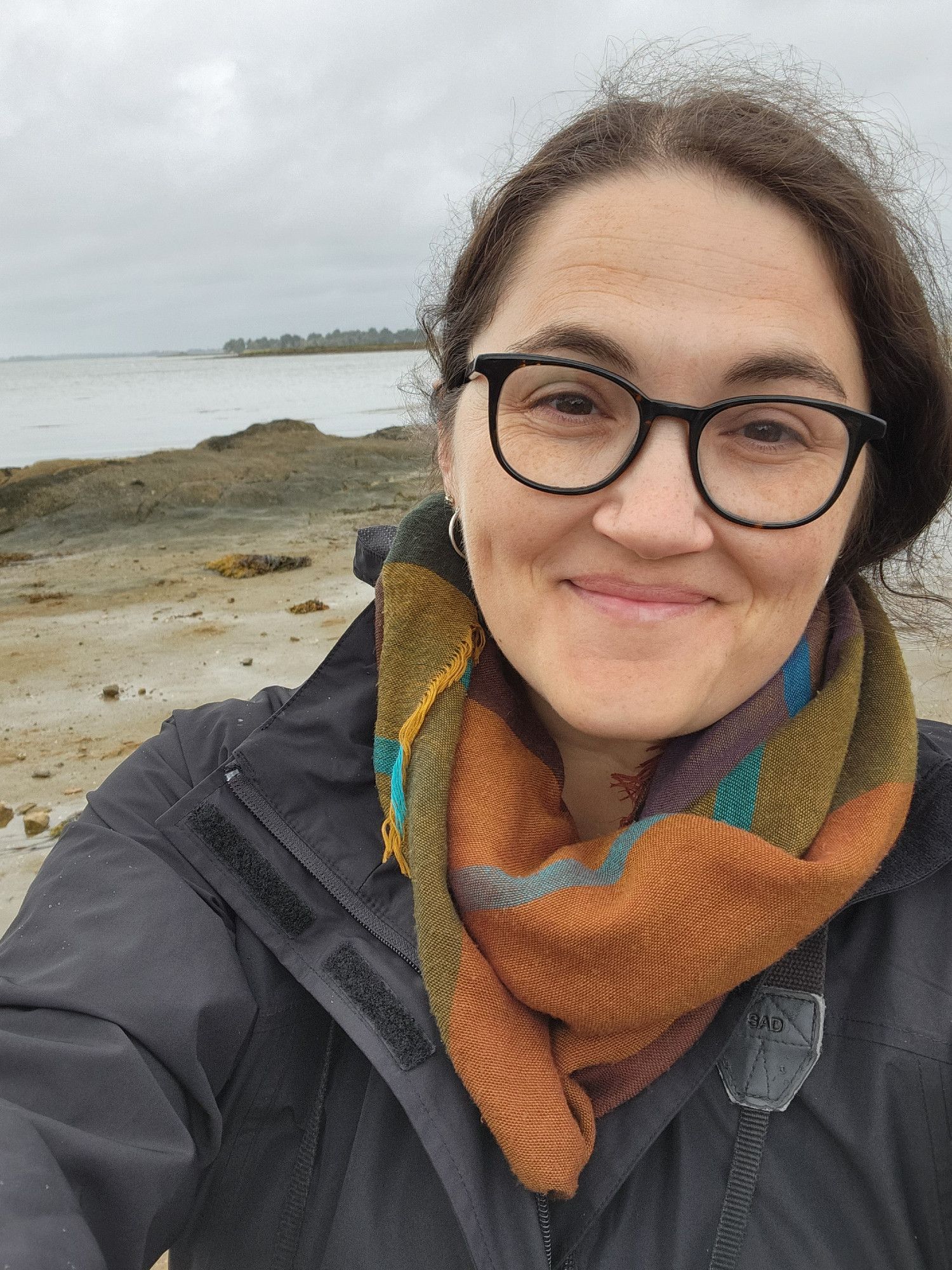 A smiling selfie of a white lady with brown hair, big glasses, a colorful scarf, and a black raincoat in front of some rocks on the shoreline