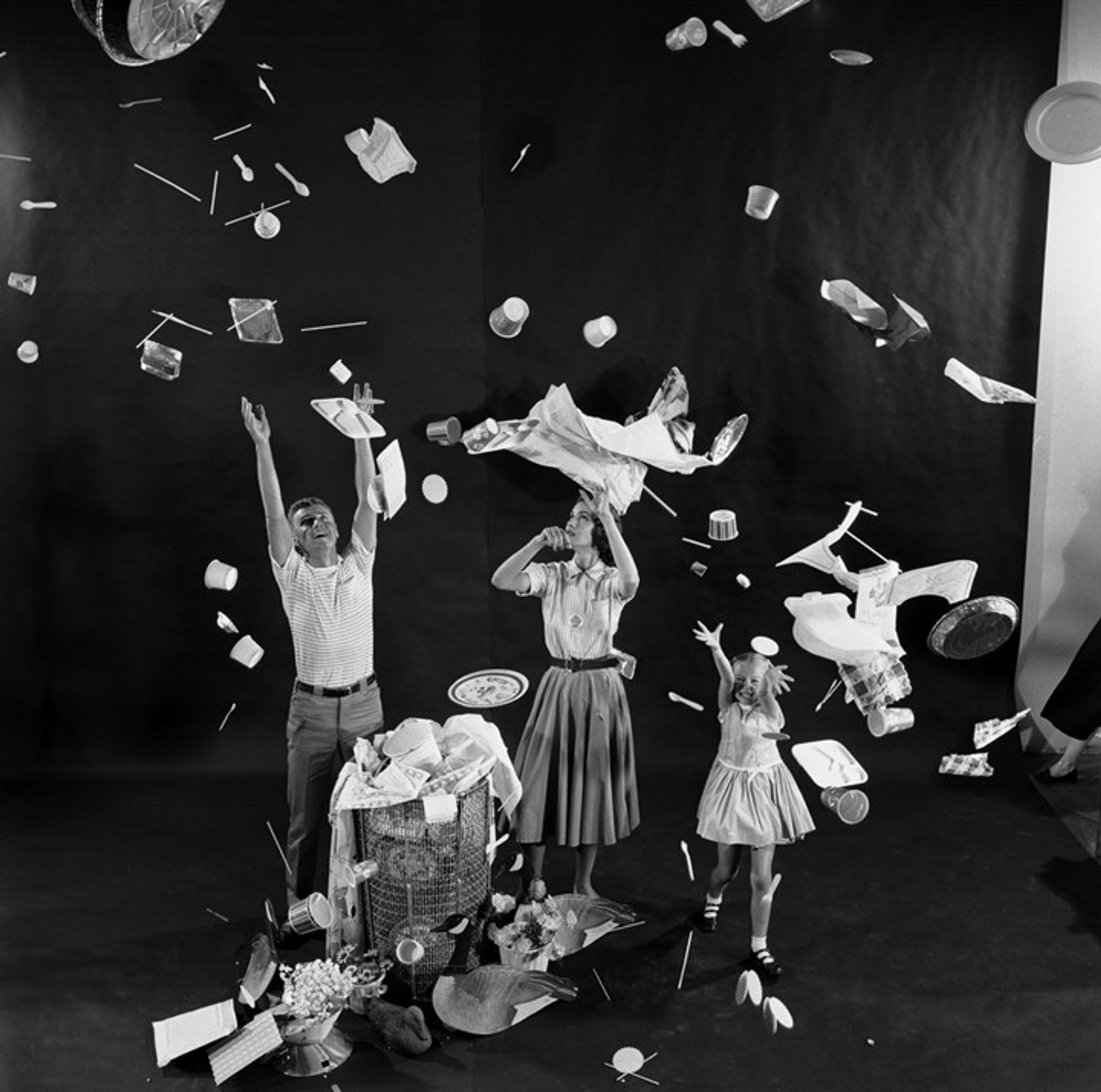 A black and white photo of a family next to an overflowing garbage can with thrown out plastic items swirling all around them - the American dream