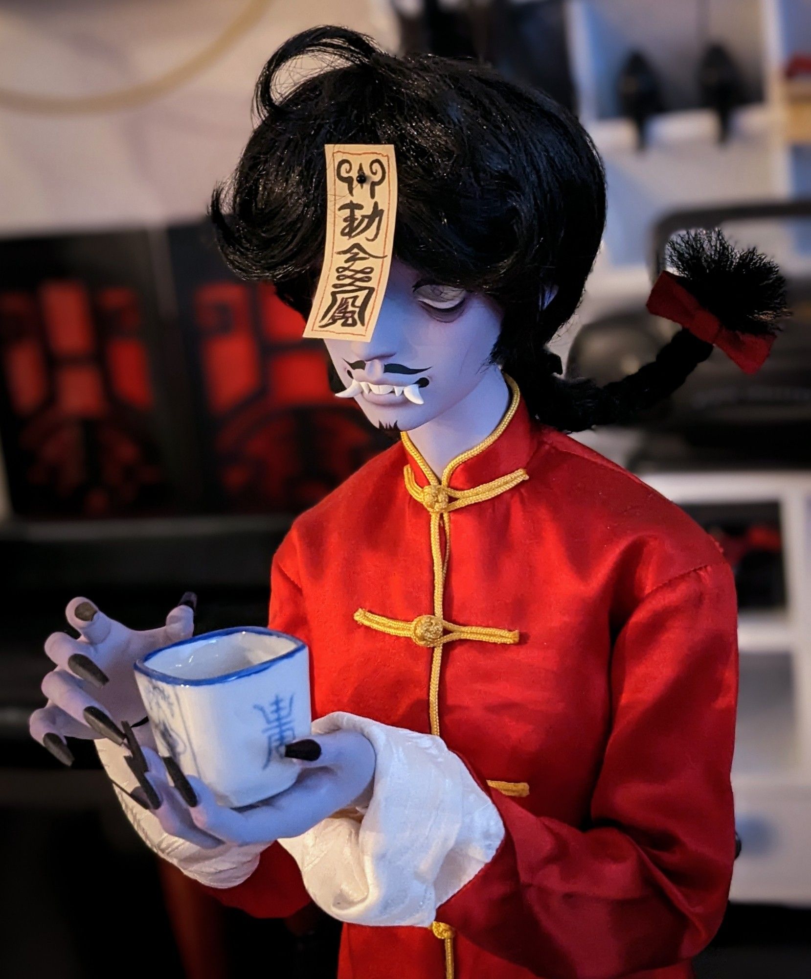 a photo of Yun holding a Chinese style tea cup

He is a Chinese hopping vampire, or 僵尸. His skin color is a ghostly  purple hue. He is wearing a red tang suit with gold duck buttons and trim. His hair is black and messy looking with a braid at the back. His eyes are sunken and deathly, his exposed teeth have sharp protruding fangs, and a mustache and small beard adorn his face. He has a magical talisman attached to his forehead to keep him docile.