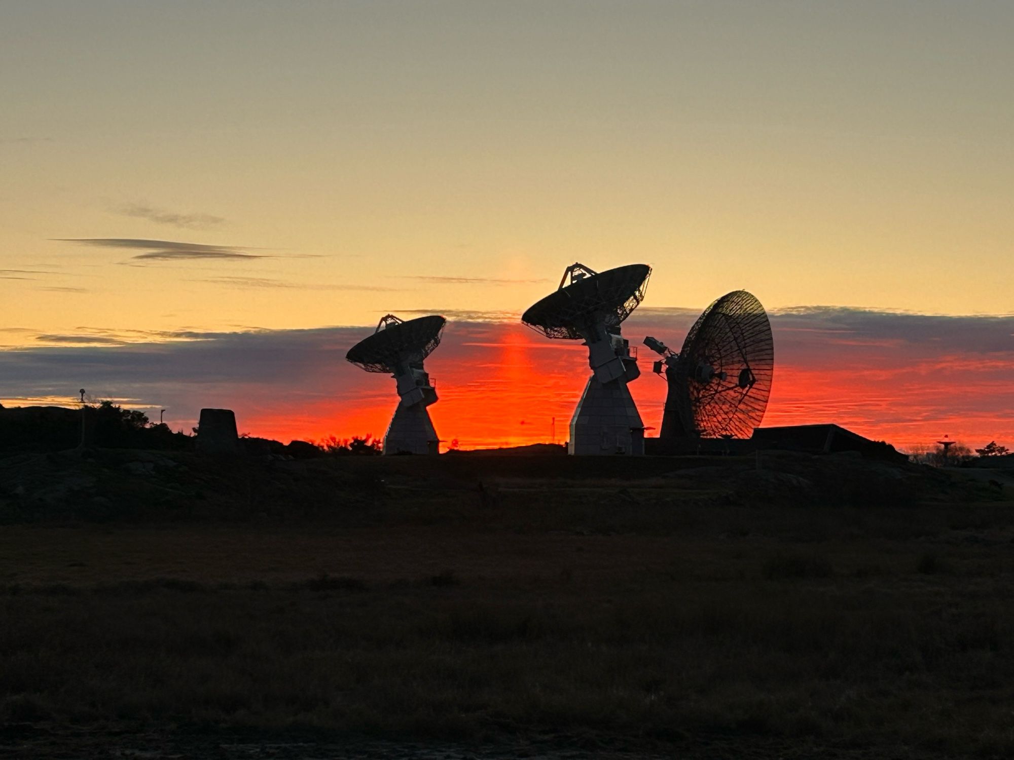 Zoomed in to three telescopes in silhouette against the sunset. The sky is clear above, but the clouds on the horizon are deep red.