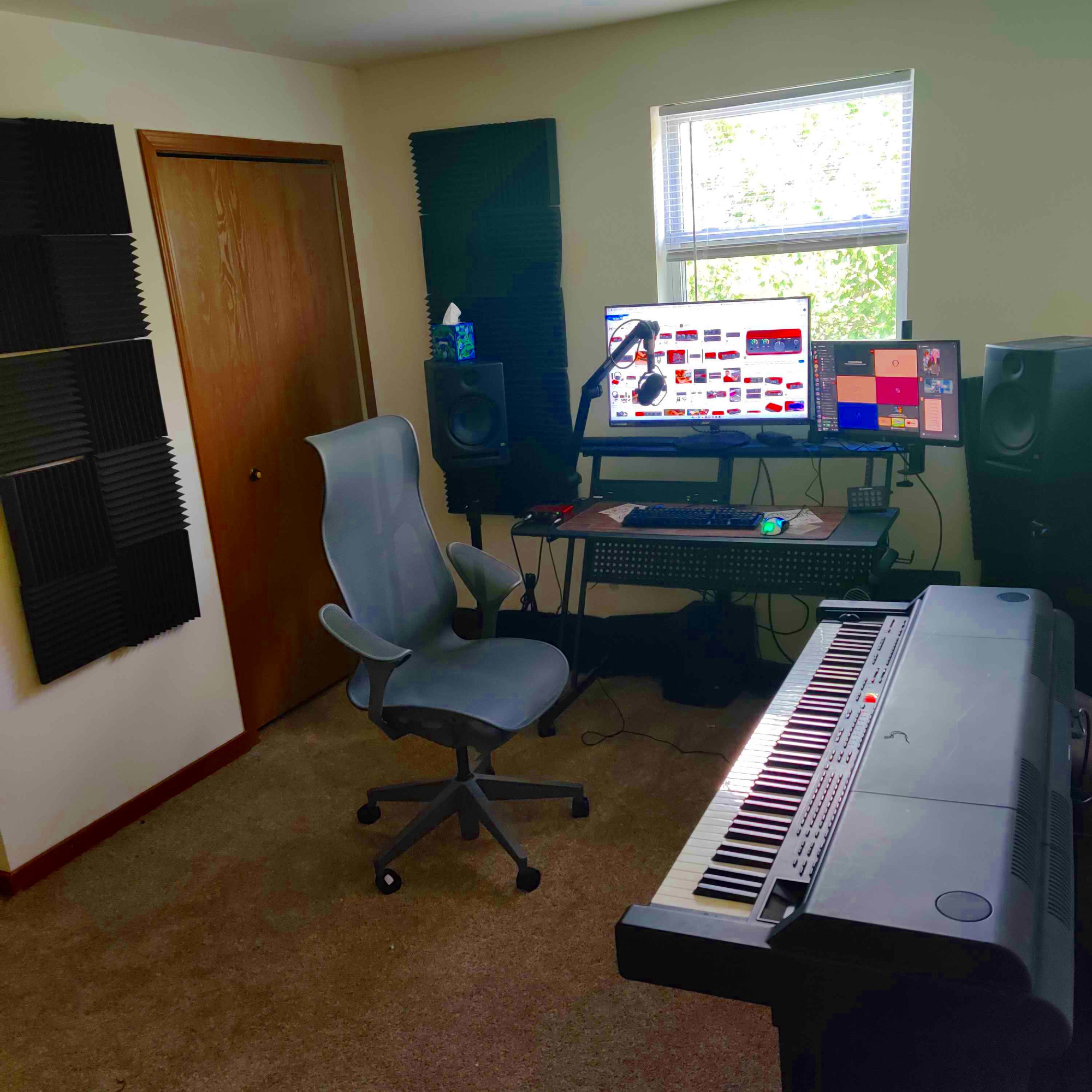 My music setup. A desk on the far side of the room against a wall with parallel speakers and a window behind it. A piano on the right and a computer chair in the center of the room facing it. On the walls are poorly hung foam panels in a checkerboard pattern.