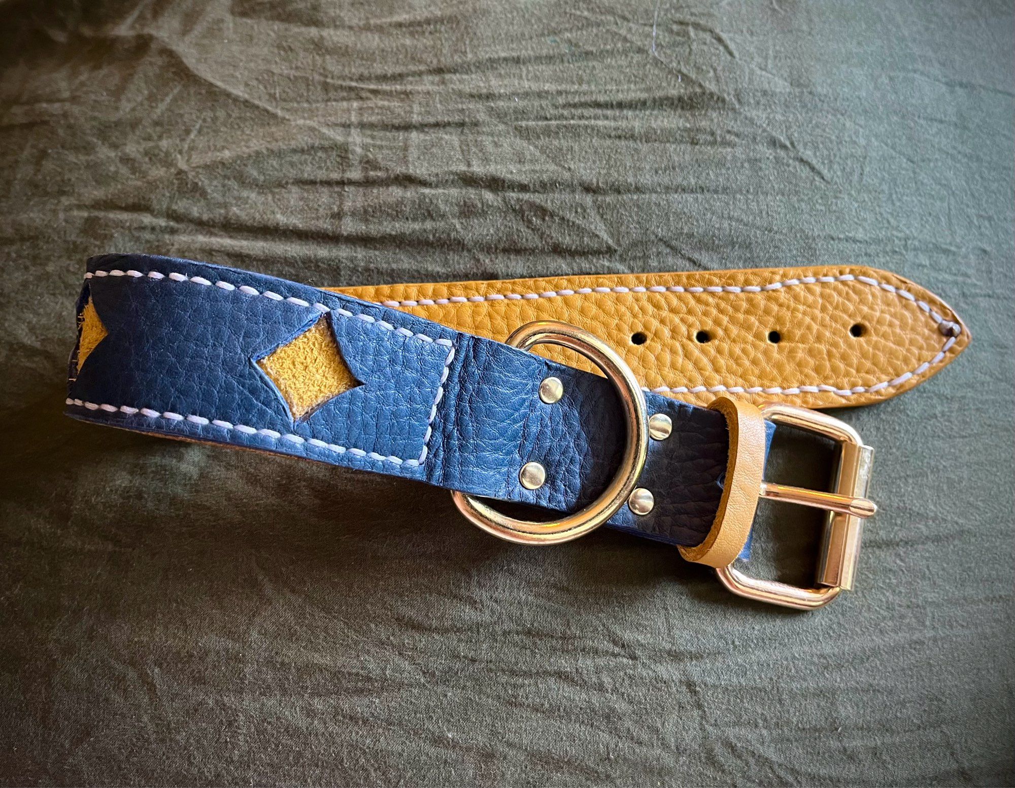 A photo of a leather collar. It is dusty blue with diamonds cut out so the yellow back piece shows through as stars. The collar has white stitching on the edges and brass hardware.