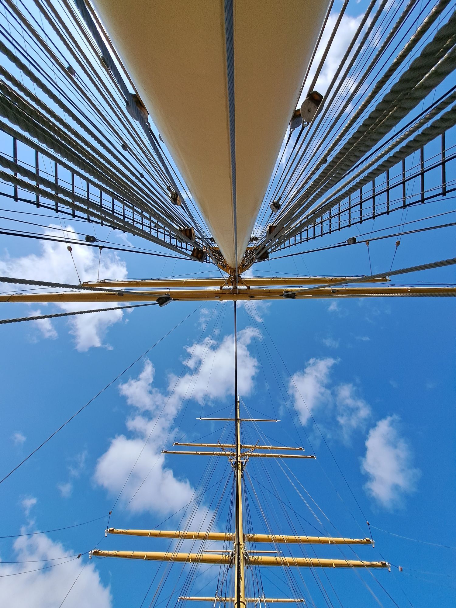 Blick gerade am Mast eines Rahseglers empor in Zentralperspektive. Unten im Bild ist ein weiterer Mast zu sehen, im Hintergrund der blaue Himmel mit wenigen Wolken.