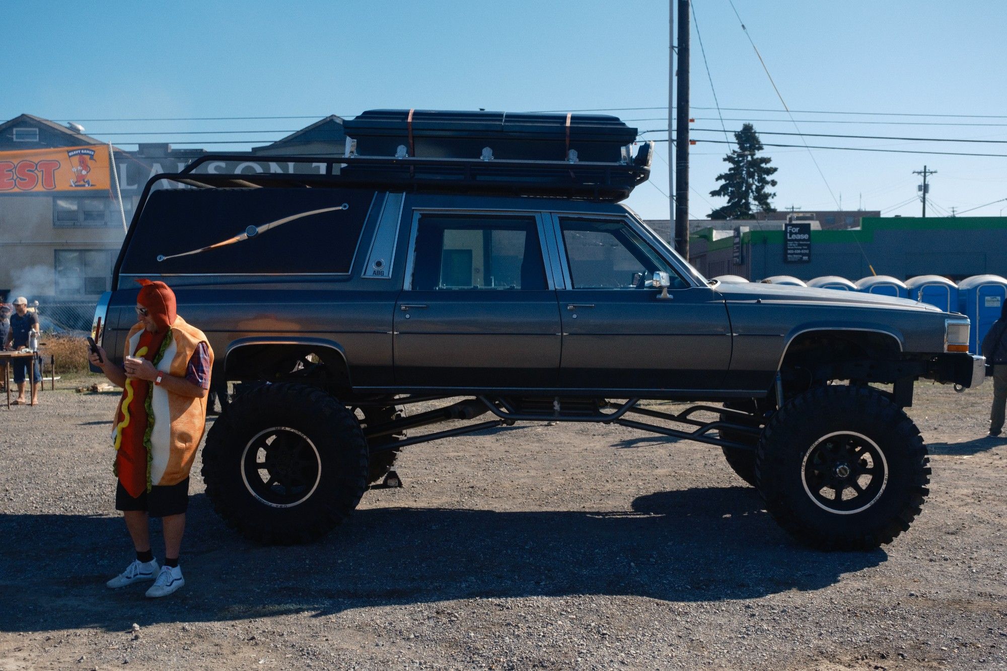 A man wearing a sausage costume looking at his phone while standing in front of a hearse with lifted suspension.