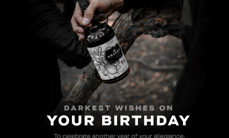A moody forest scene: someone holds a bottle of Kraken Black Spiced Rum, the label showing its iconic sea monster. The lighting is dim, casting the bottle in sharp relief against a gnarled tree branch. Text over the image reads: “Darkest wishes on your birthday. To celebrate another year of your allegiance.”