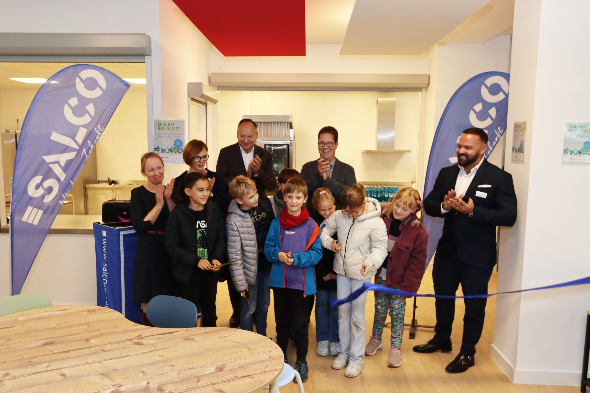 Kinder der Wallschule schneiden das symbolische Band für die Eröffnung der neuen Mensa durch. Daneben stehen Schulleiterin Anja Knauß, ihre Stellvertreterin Sandra Spengel, Landrat Oliver Quilling, Bürgermeister Jan Werner sowie Salco-Geschäftsführer Adrian Khalifé. 