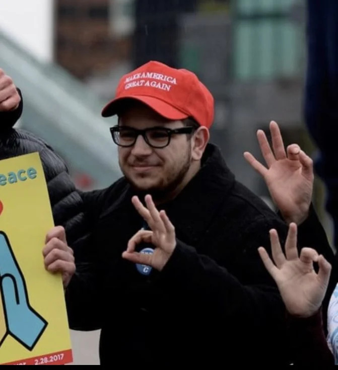 A man wearing a MAGA hat and making a white-power hand gesture