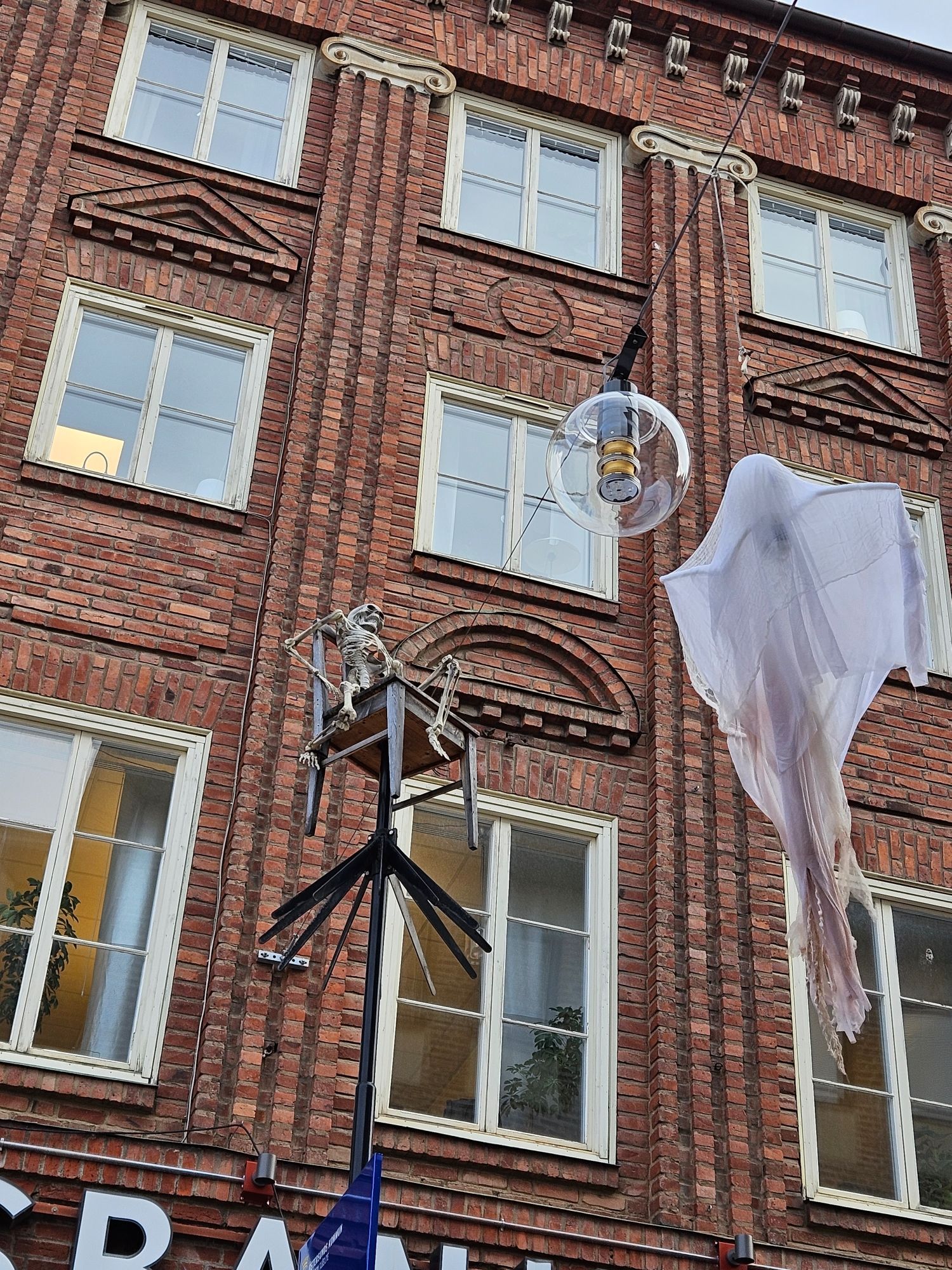 Halloween decoration skeleton chills out on a chair mounted on a light post while a ghost hangs from a line across from it