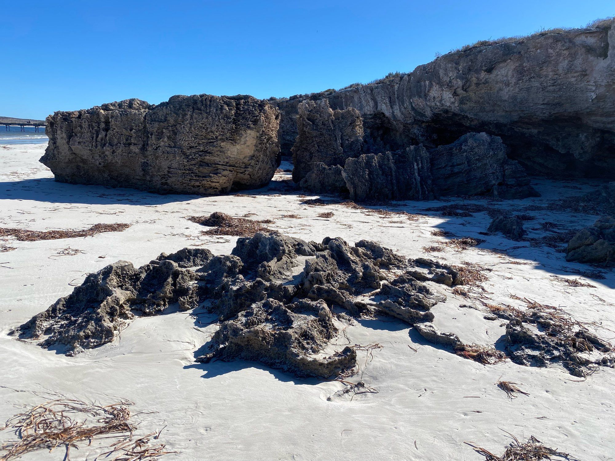 A rock formation with a small arch caused by erosion sitting in white sand with blue sky above.