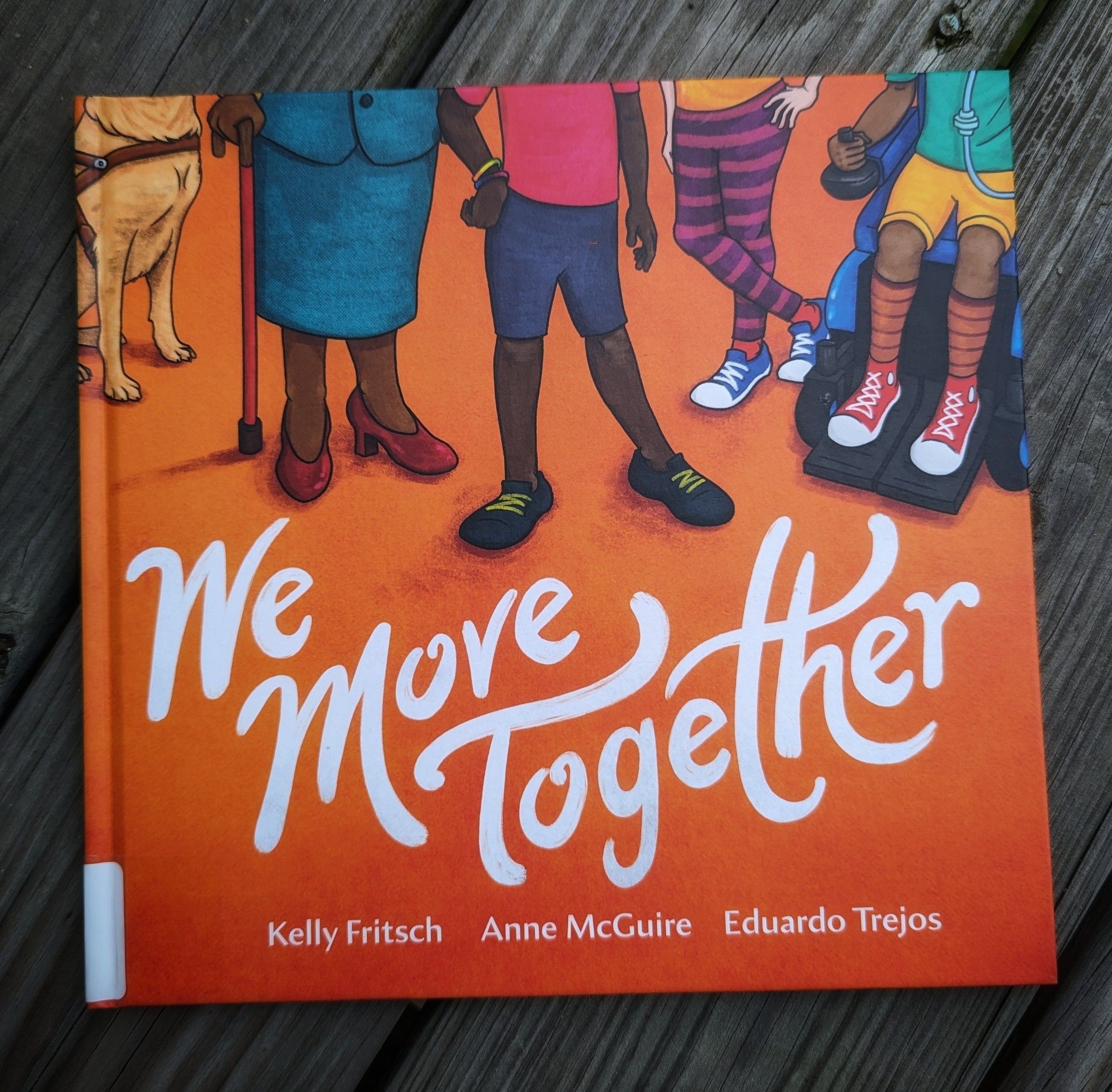 Photo of the #PictureBook: We Move Together by Kelly Fritsch, Anne McGuire, & Eduardo Trejos. The cover is a deep orange with white lettering. The top half of the book shows the lower half of 4 humans of varying skin tones and a dog with a harness that suggests it's an assistance animal.  One human holds a cane, and another is using a wheelchair.