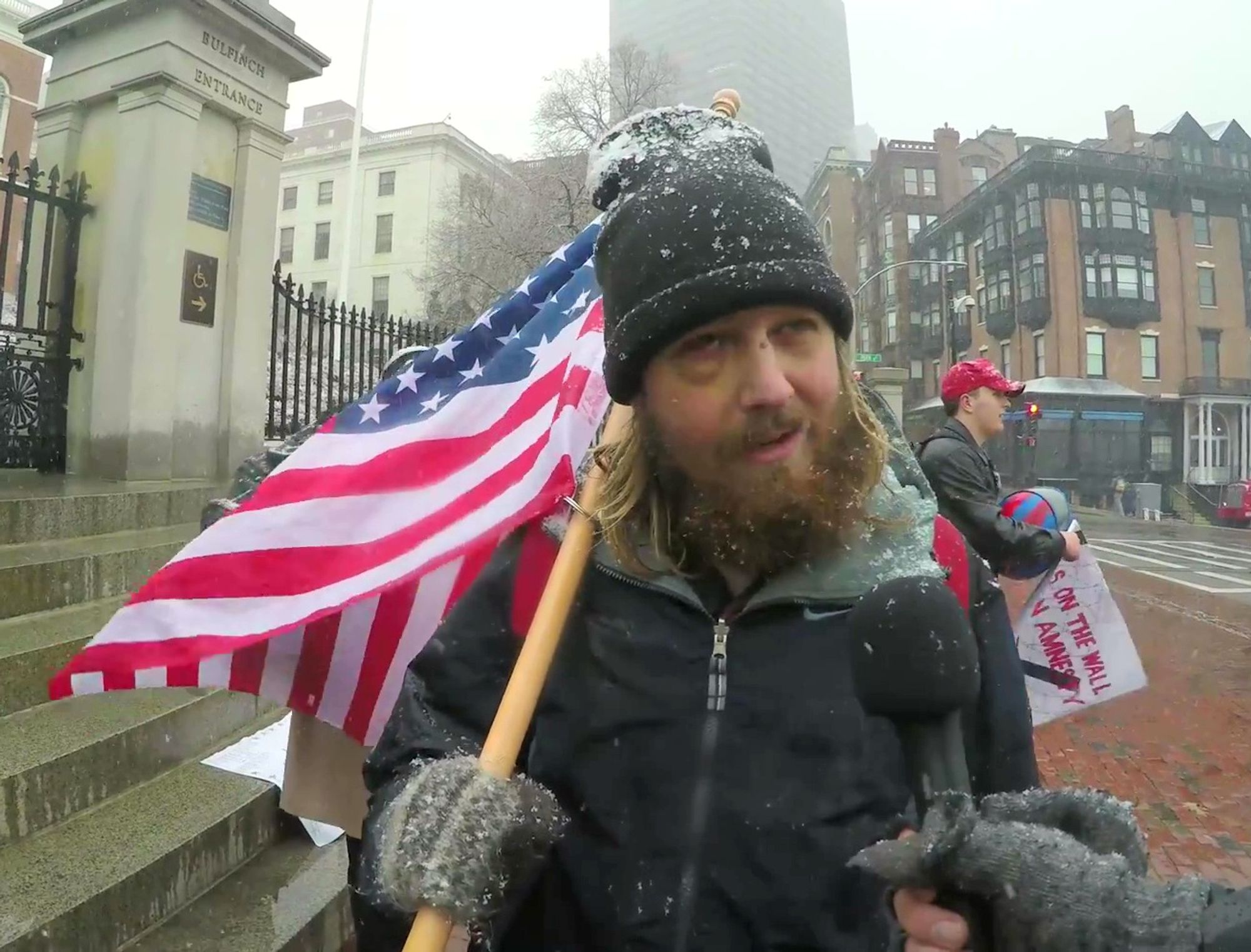 A still of video. It depicts Jarek Reihner at the steps outside the front gate of the Massachusetts State House during December 2017. Reihner is dressed in a black winter jacket, gray knit mittens, and a tall black beanie. His clothes, particularly his beanie, have wet flakes of snow clinging to them. He carries an American flag over his right shoulder. His hair is long and blond, and his beard is puffy and unkempt. His right eye is swollen. He is being interviewed by an unseen white man holding a mic. Behind Reihner, Boston is slightly foggy. The stone staircase in front of the State House, brick sidewalk, and road nearby are visibly dampened from previously melted snow.