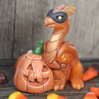 An orange dragon figure with a black mask sitting by a toothy smiling jack o lantern. He is surrounded by candy corn. 