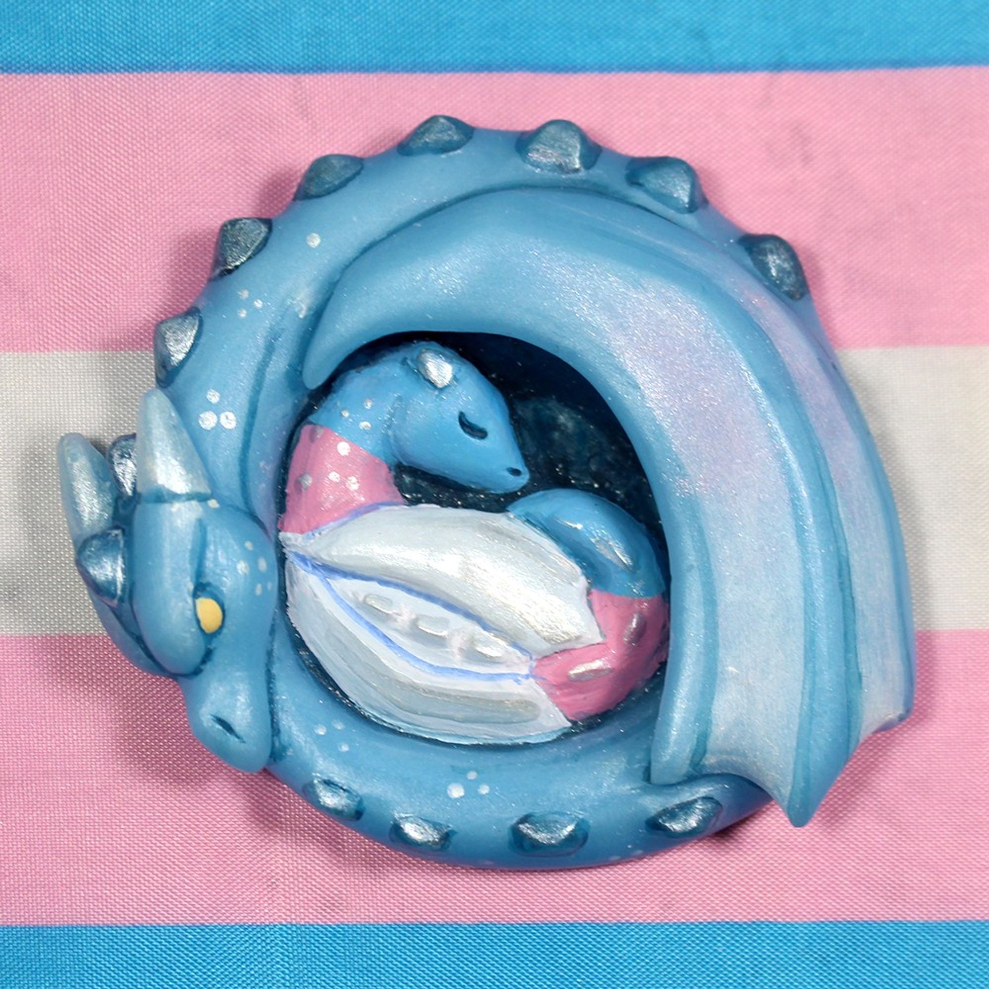 A light blue dragon curls around a baby dragon colored like the trans pride flag.  In the background is the trans pride flag.