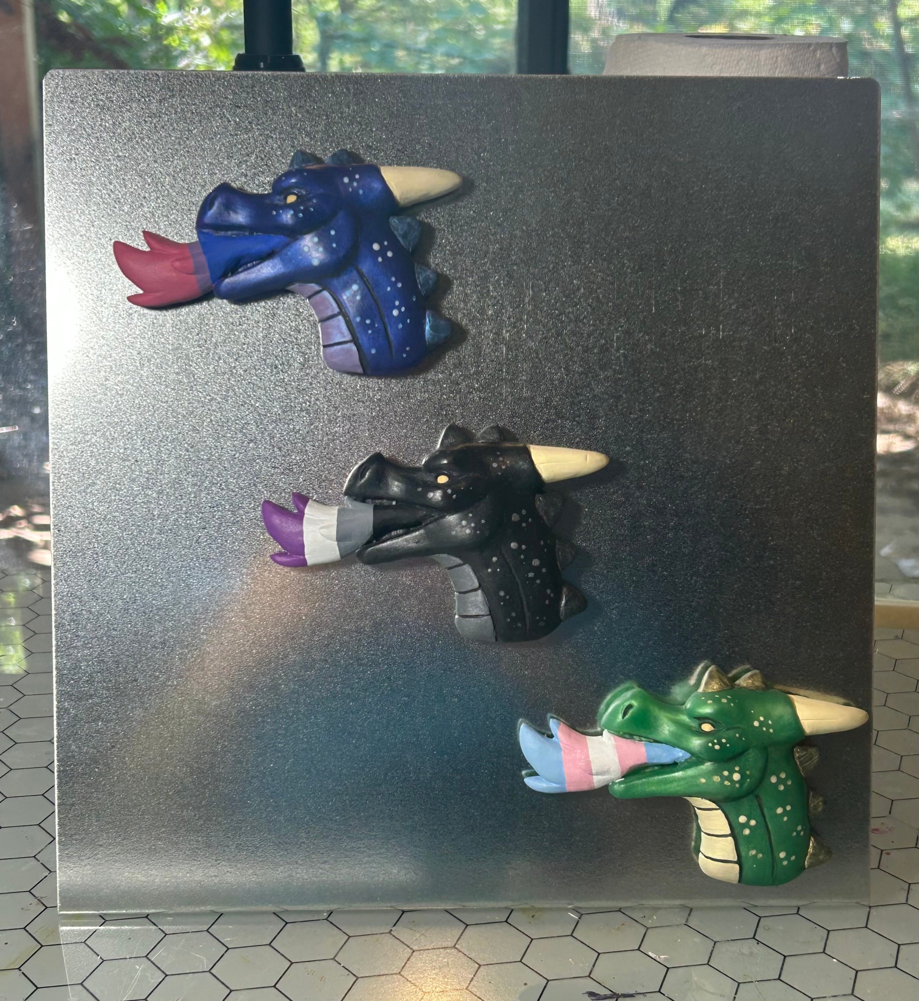 A silver magnet board displaying three fire breathing dragon busts. They are different colors with Pride flag themed flames. 
