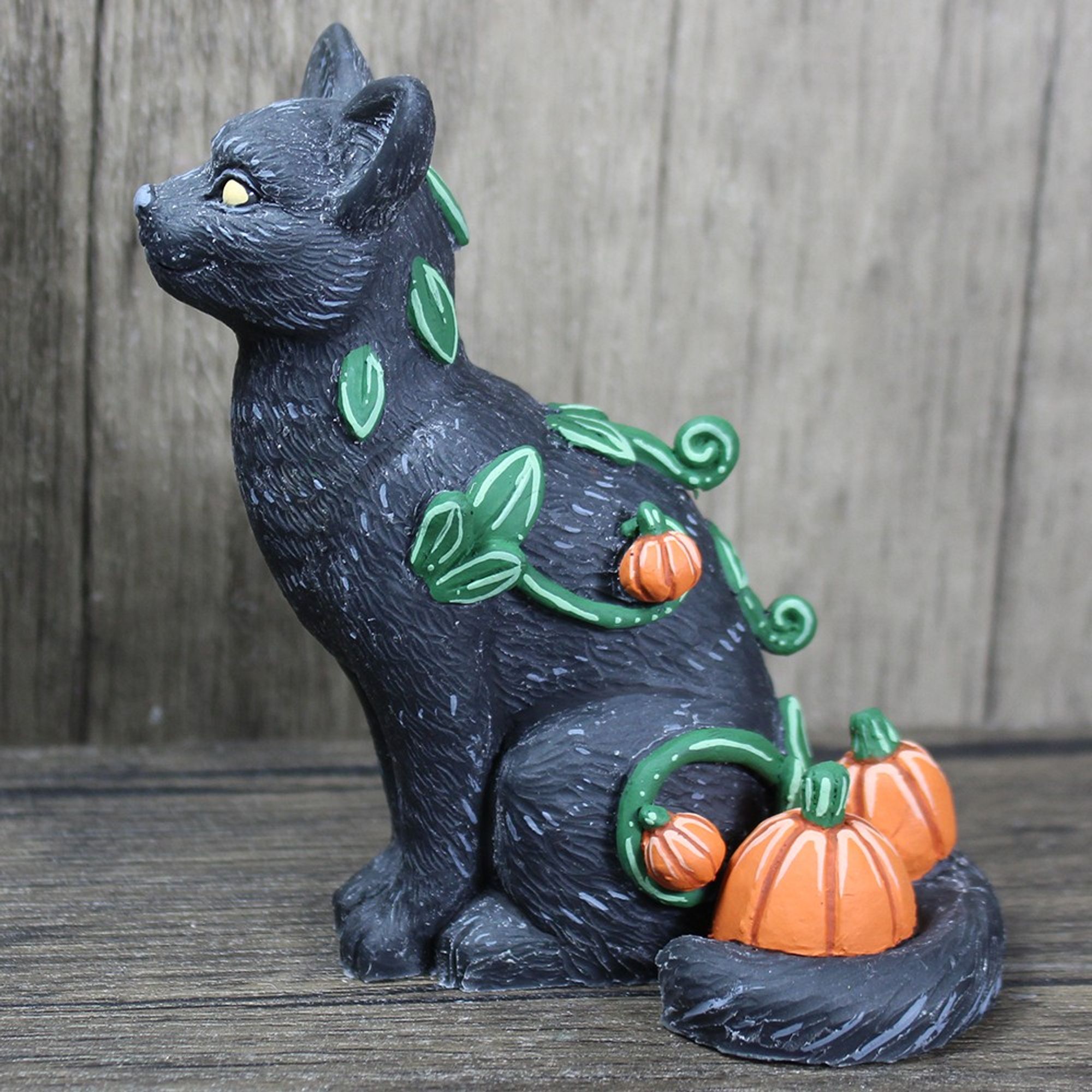 A figure of a black cat sitting.  It's tail is curled around a pair of pumpkins.  On it's body there are vines and leaves and miniature pumpkins.