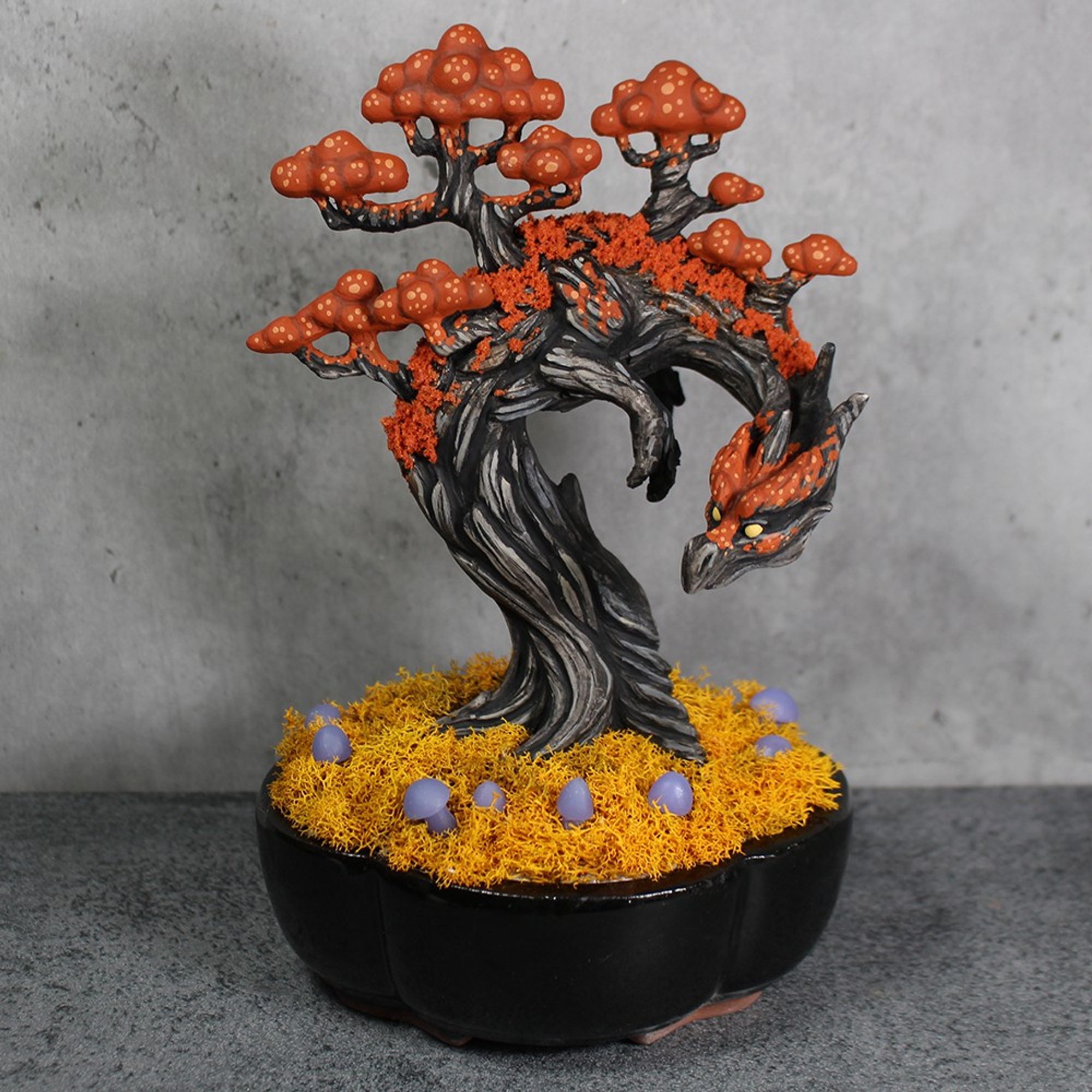 An orange and black tree dragon planted in a black bonsai pot.  He is surrounded by golden moss and purple mushrooms.