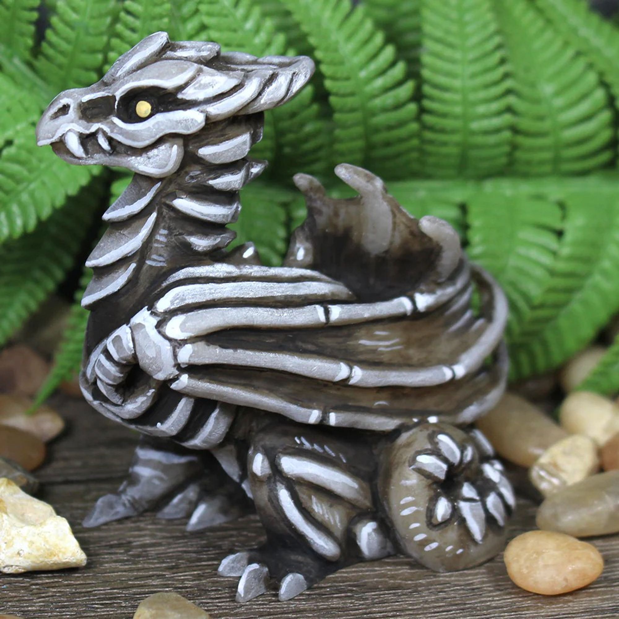 A  black and white skeleton dragon figure.  He has yellow eyes and is sitting amongst pebbles and ferns.