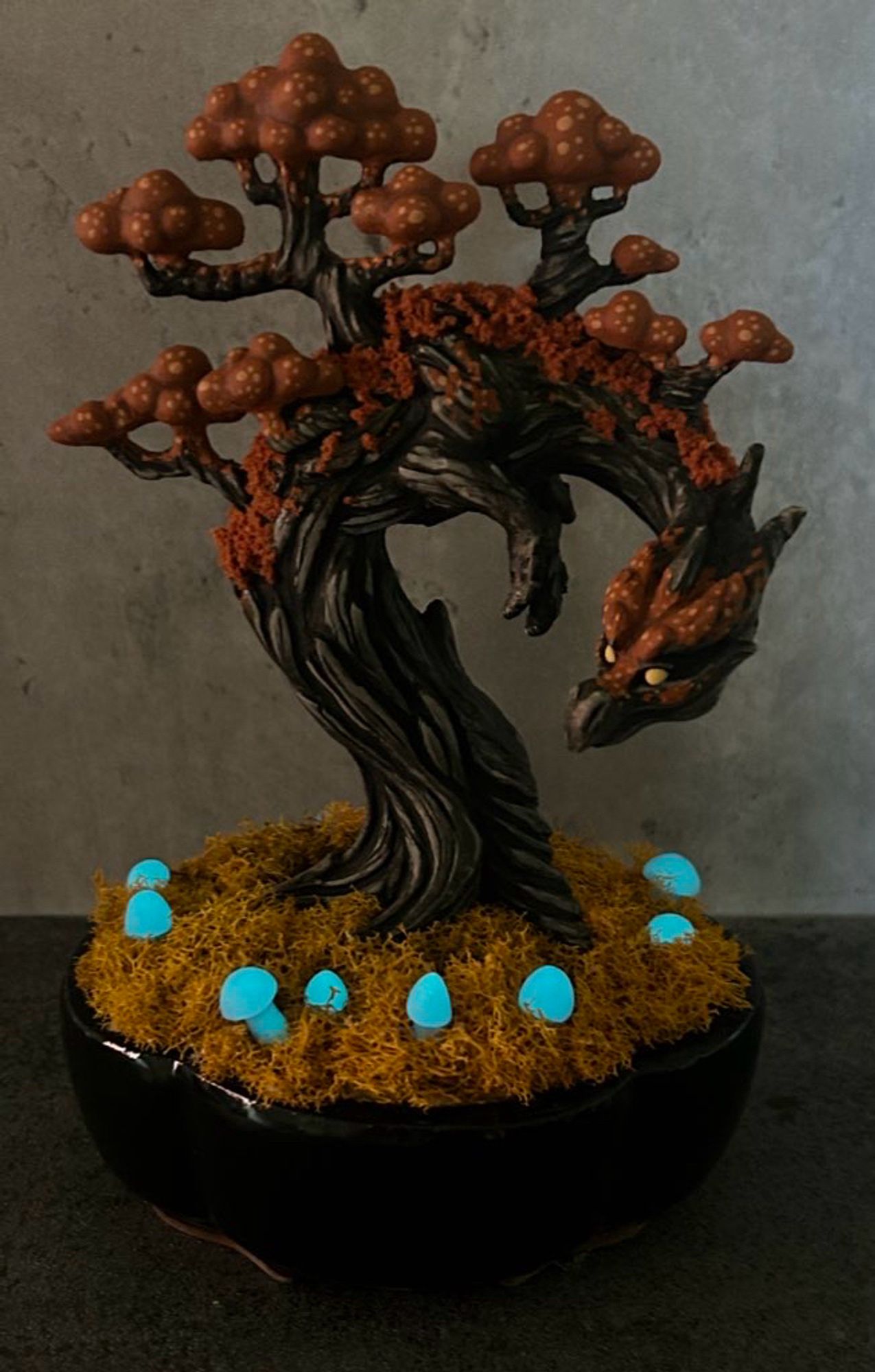 A black and orange Halloween themed tree dragon planted in a black pot surrounded by golden moss and glowing blue mushrooms.