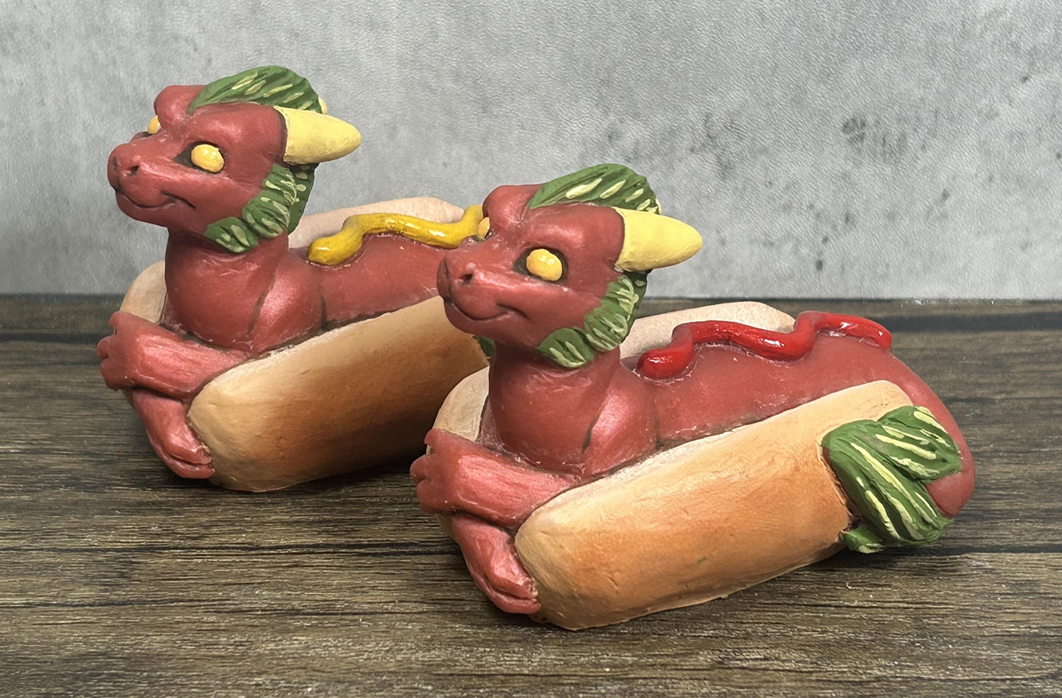 Two figures of hotdog themed dragons sitting in buns.  They have green manes and tails that resemble relish with squirts of mustard and ketchup on their backs.