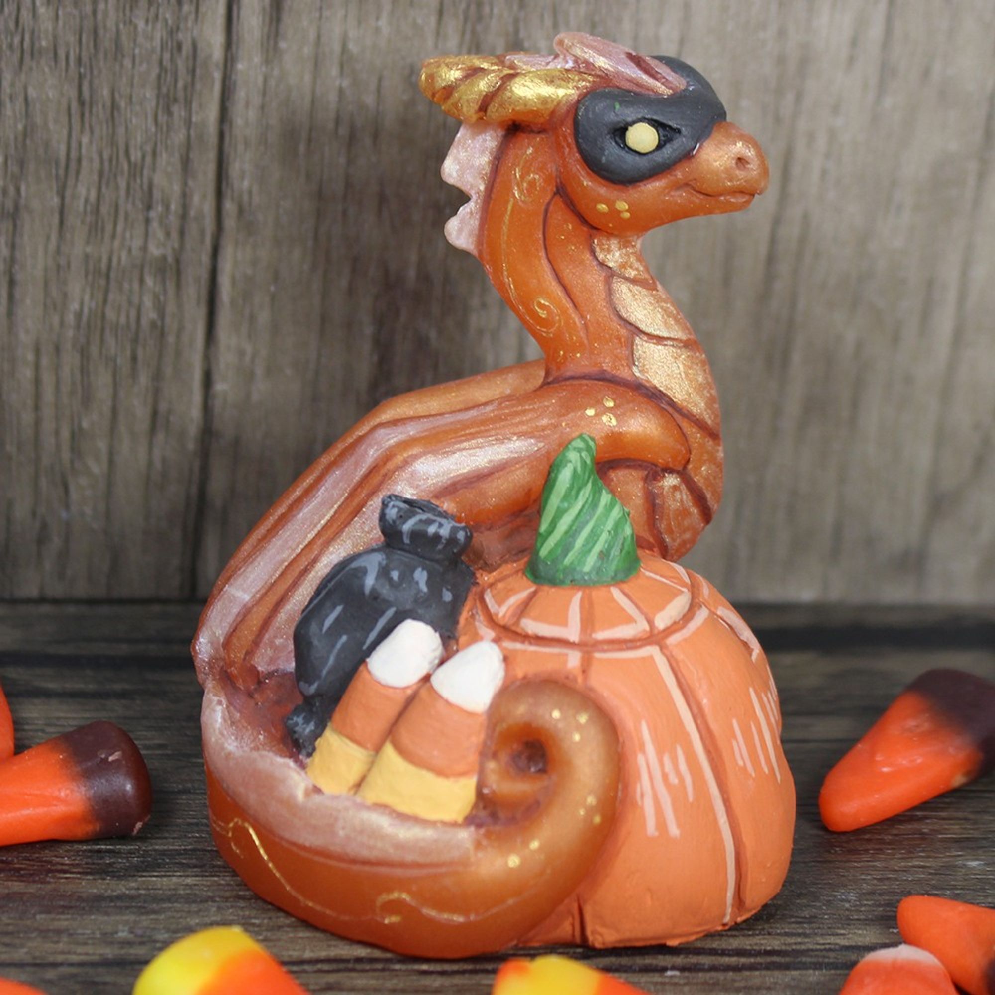 An orange dragon curled around a jack-o-lantern and some candies.  He is wearing a black mask and is surrounded by candy corn.