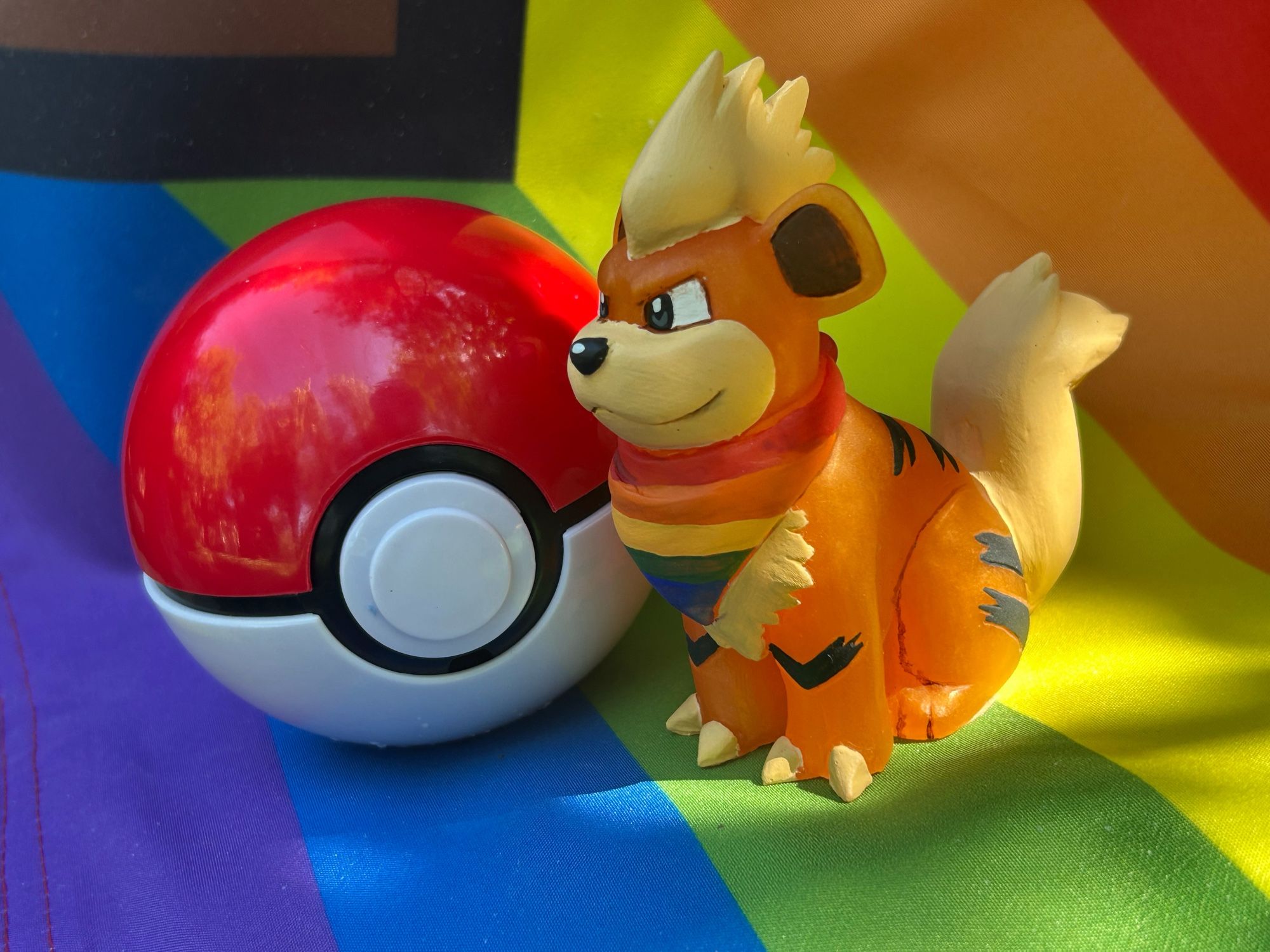 A growlithe figure wearing a rainbow bandana. He is standing next to a pokeball on a rainbow background.