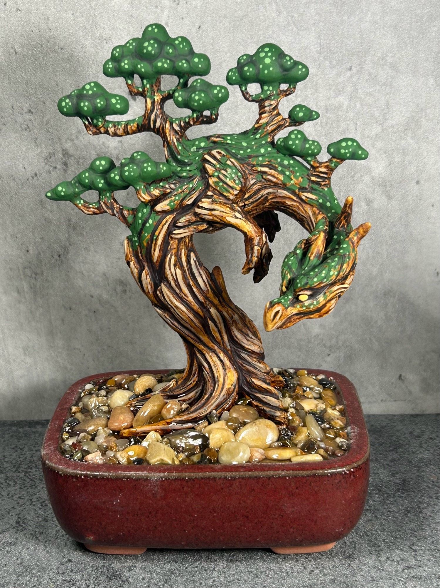 A tree dragon figure planted in a red bonsai pot. The dragons roots are surrounded by pebbled.