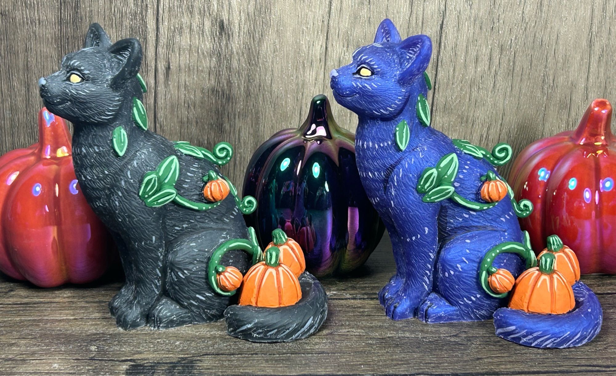 Two cat figures, one black and one purple. Their tails are curled around a pair of pumpkins. Leaves, vines and mini pumpkins grow from their fur.