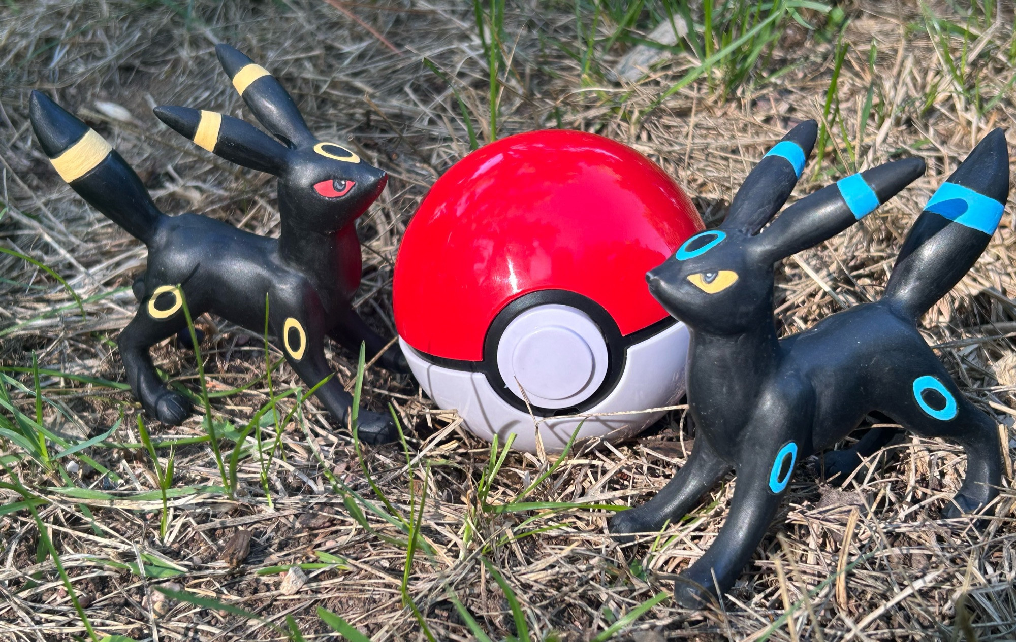 Two umbreon figures on either side of a pokeball sitting on grass.