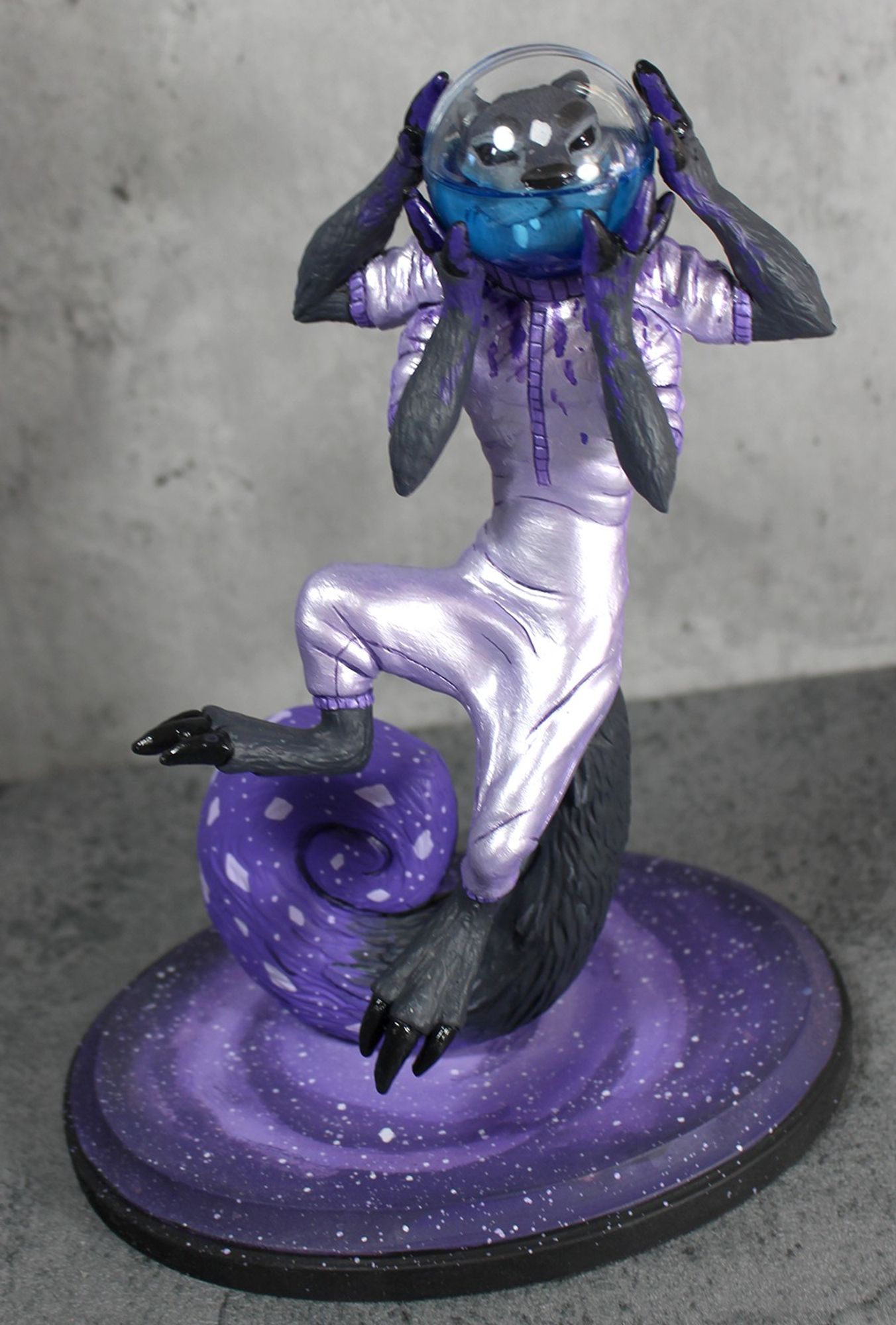 A sculpture of a space otter with four arms, hovering above a galaxy painted base.  It is colored in purples and greys.