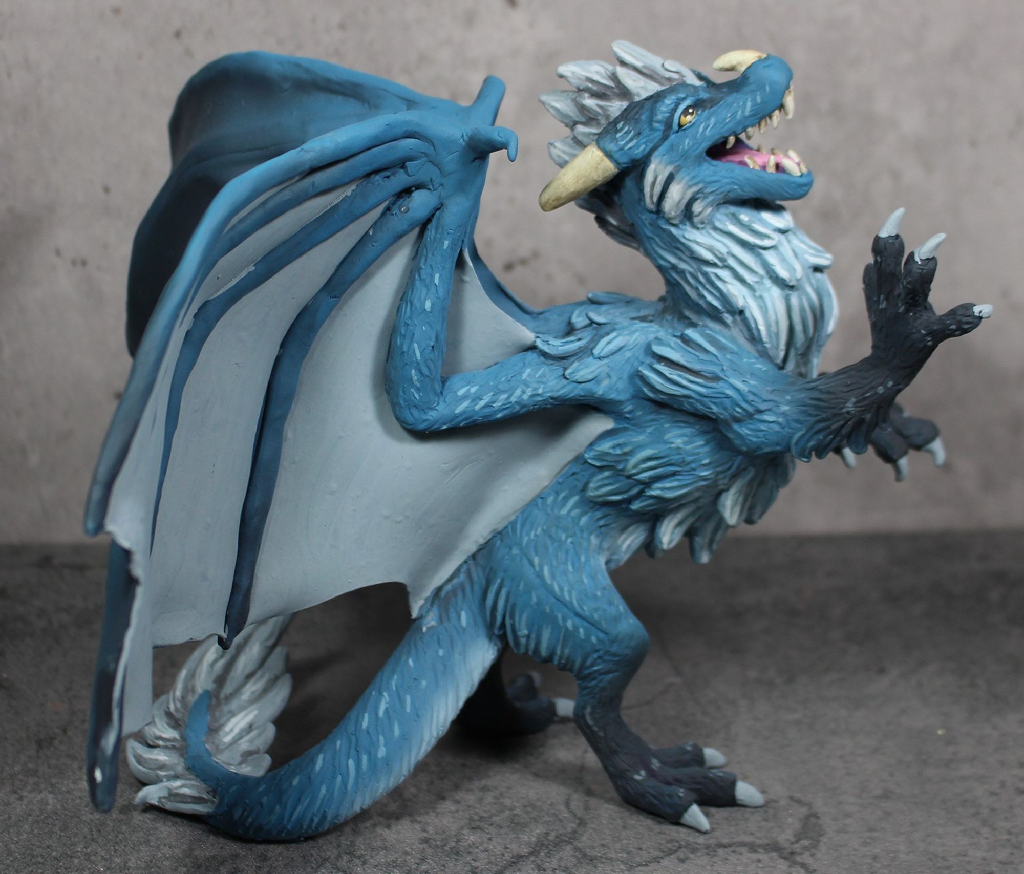 Second side view - A sculpture of a furry dragon rearing on his hind legs and pawing at the air.  He is blue with a white underside and dark tipped feet.  His mane and wings are silver.